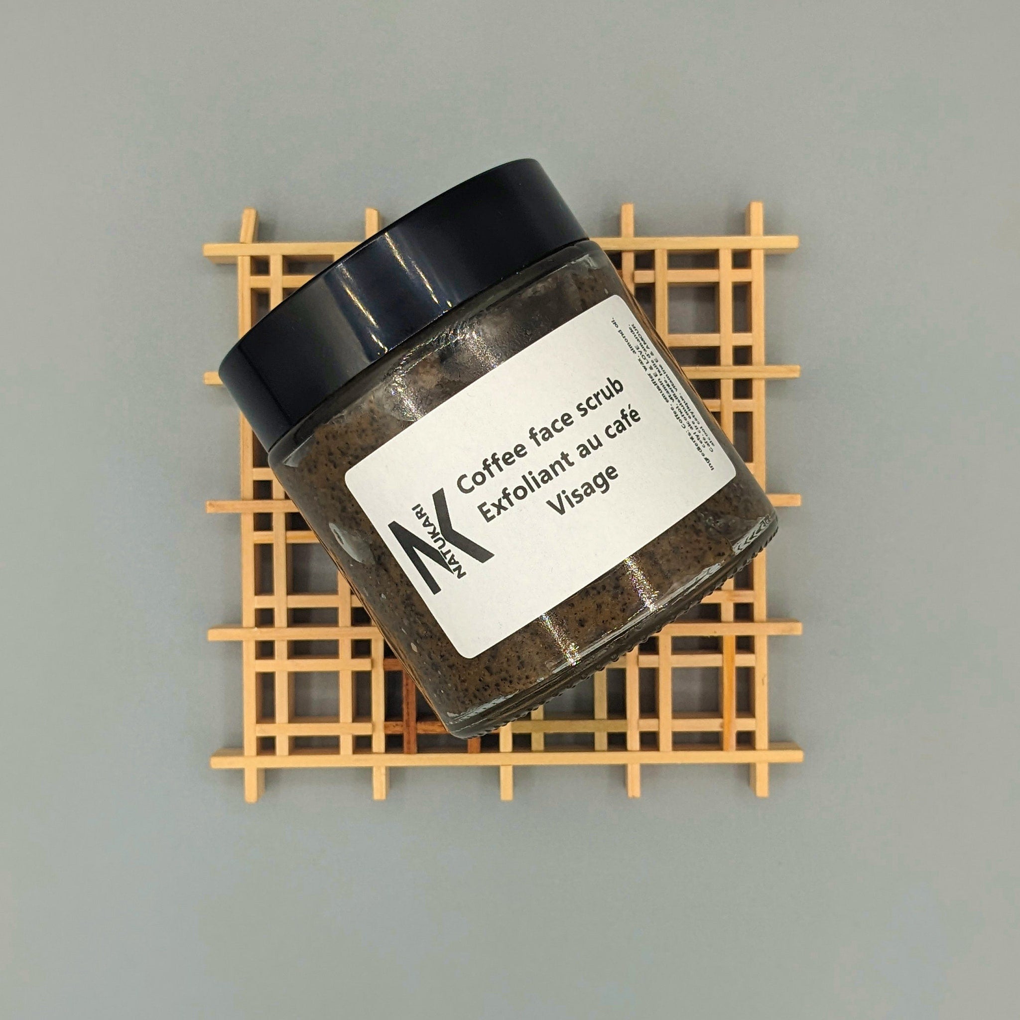 Coffee face scrub jar with black lid, brown contents, and NATUKARI label on wooden stand