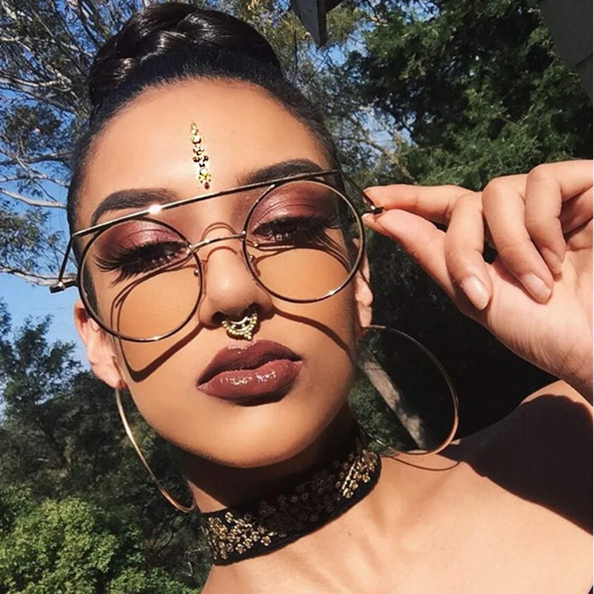 Round sunglasses with gold frame and amber lenses, worn by a person with dark makeup and jewelry