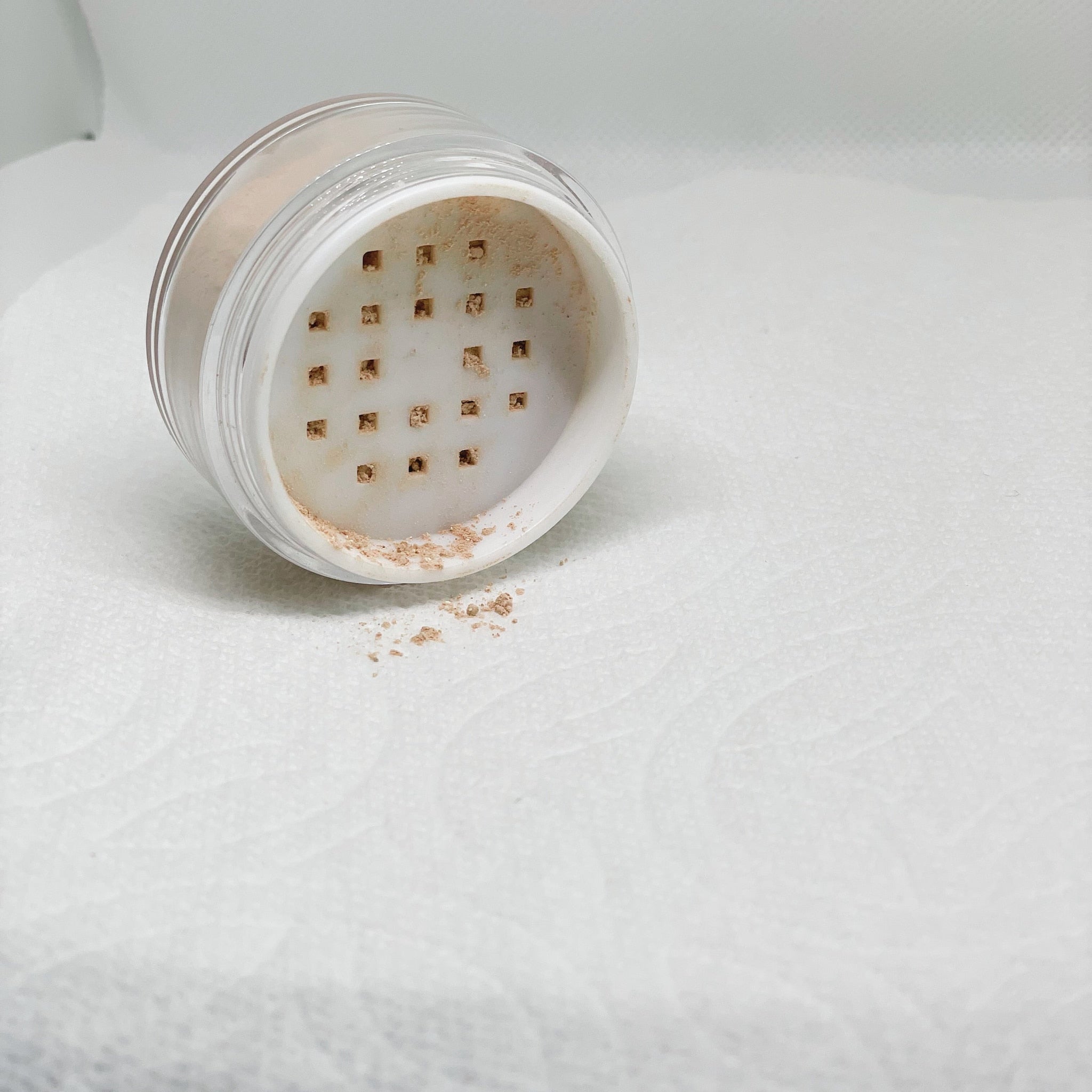 Loose powder makeup in clear plastic container with beige powder, spilled on white textured surface