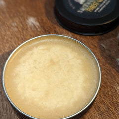 Beard balm in a silver tin with light yellow cream and black lid in background on brown surface