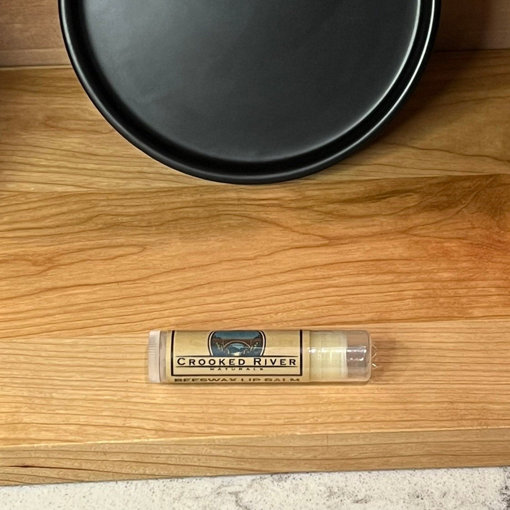 Lip balm tube with clear plastic, beige label, and "Crooked River" text on wood surface