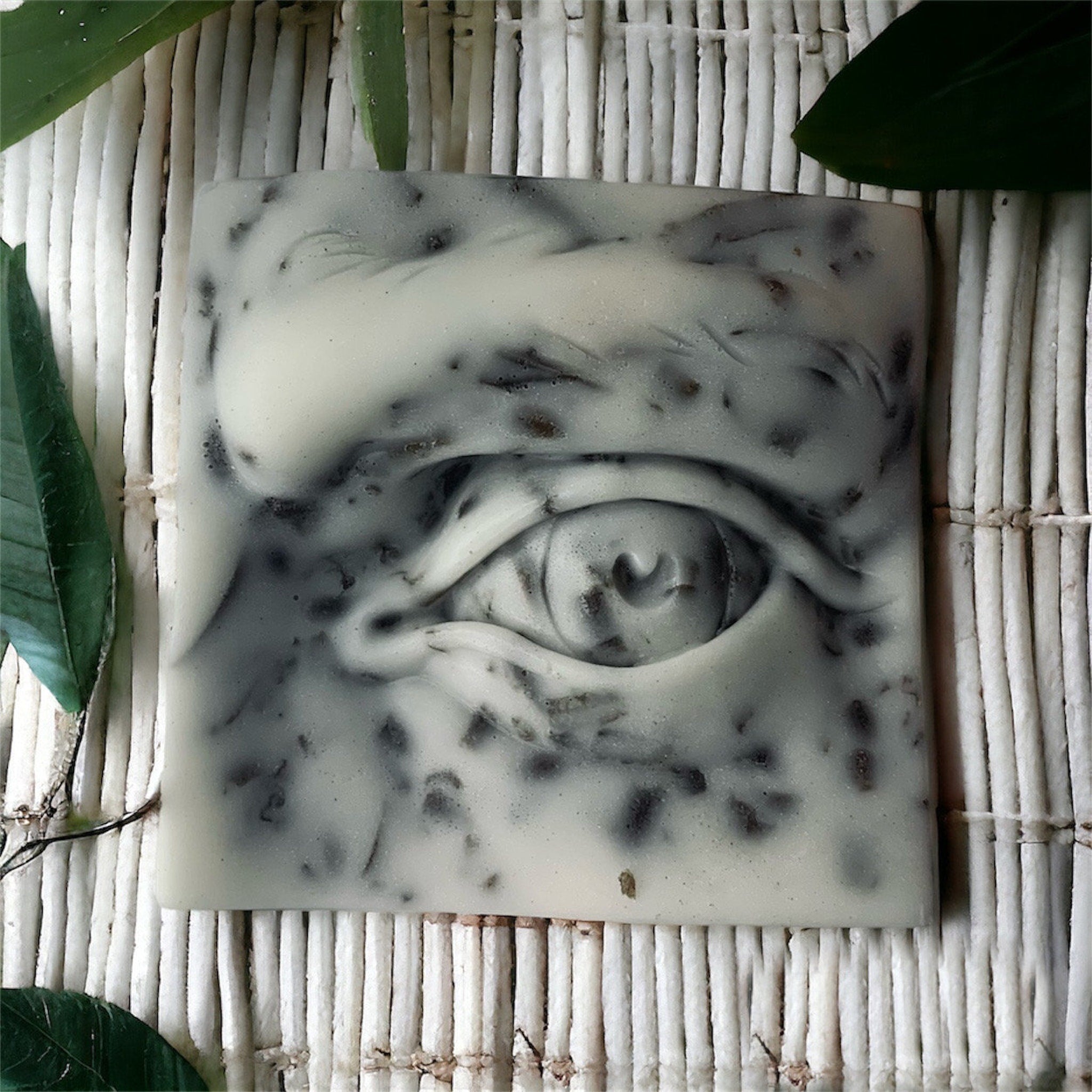 Soap bar with eye design, white and grey colors, speckled pattern on a white bamboo mat
