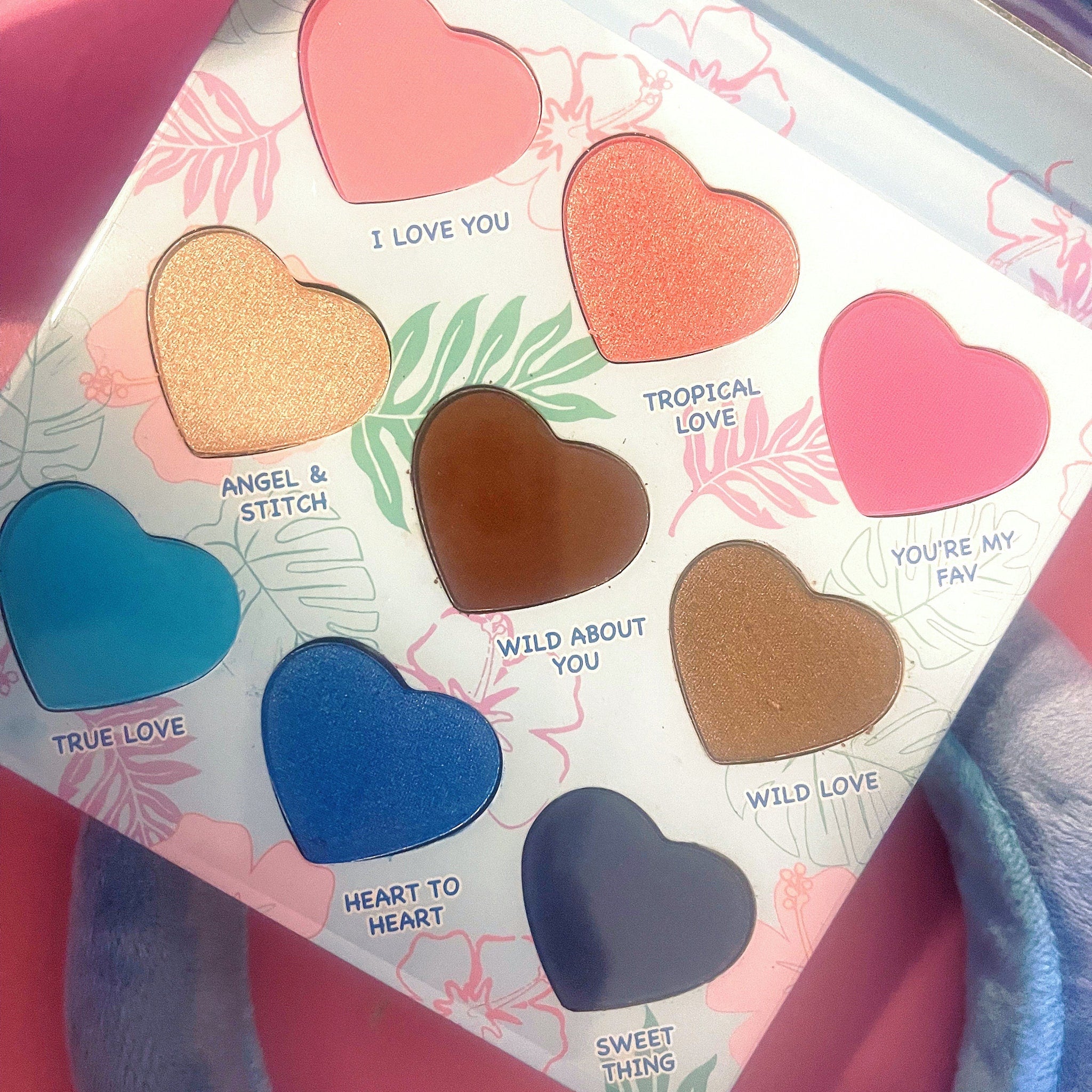 Heart-shaped eyeshadow palette with various colors and text labels, floral pattern background