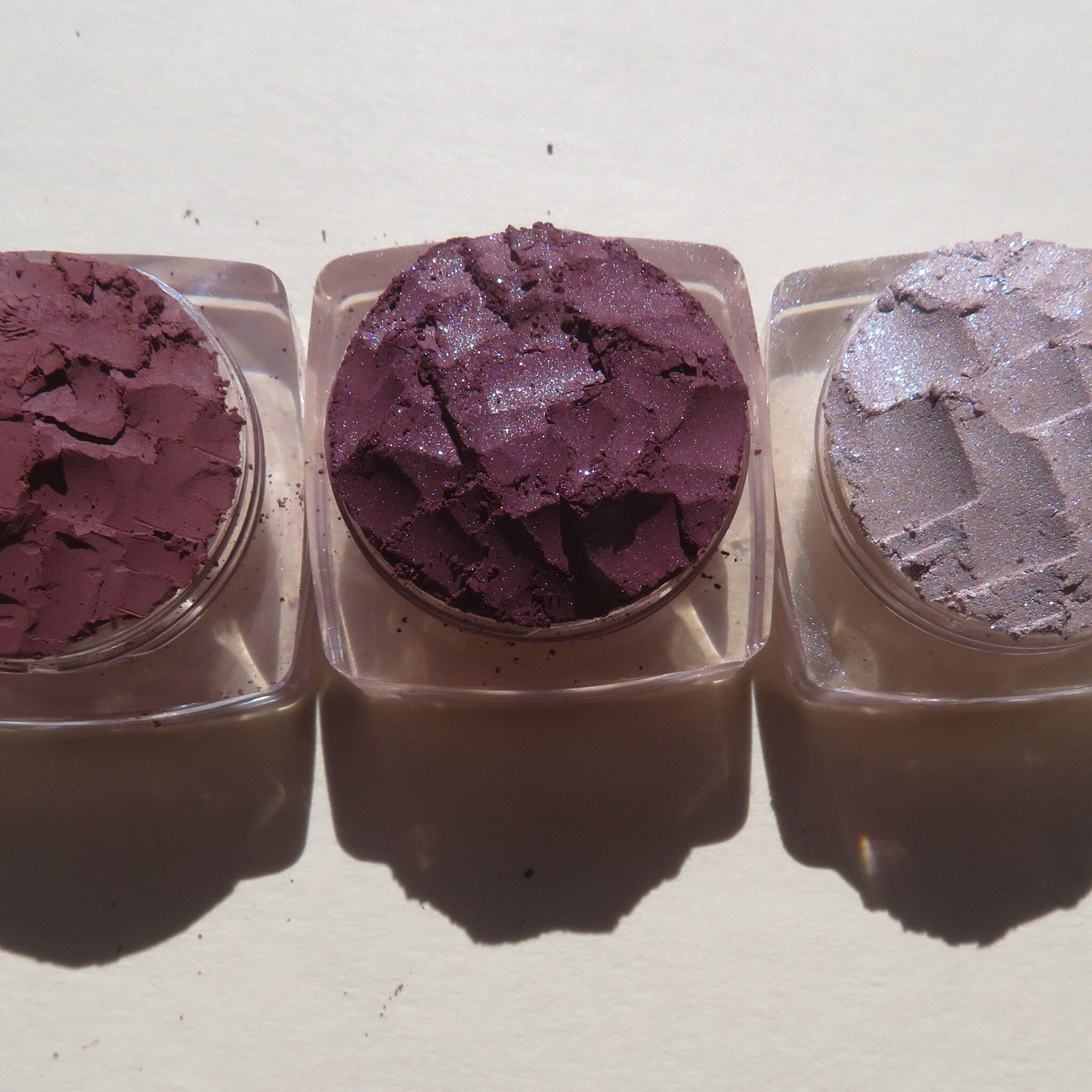 Three purple shimmer loose pigment jars with mauve matte shade, displayed on marble surface with brush