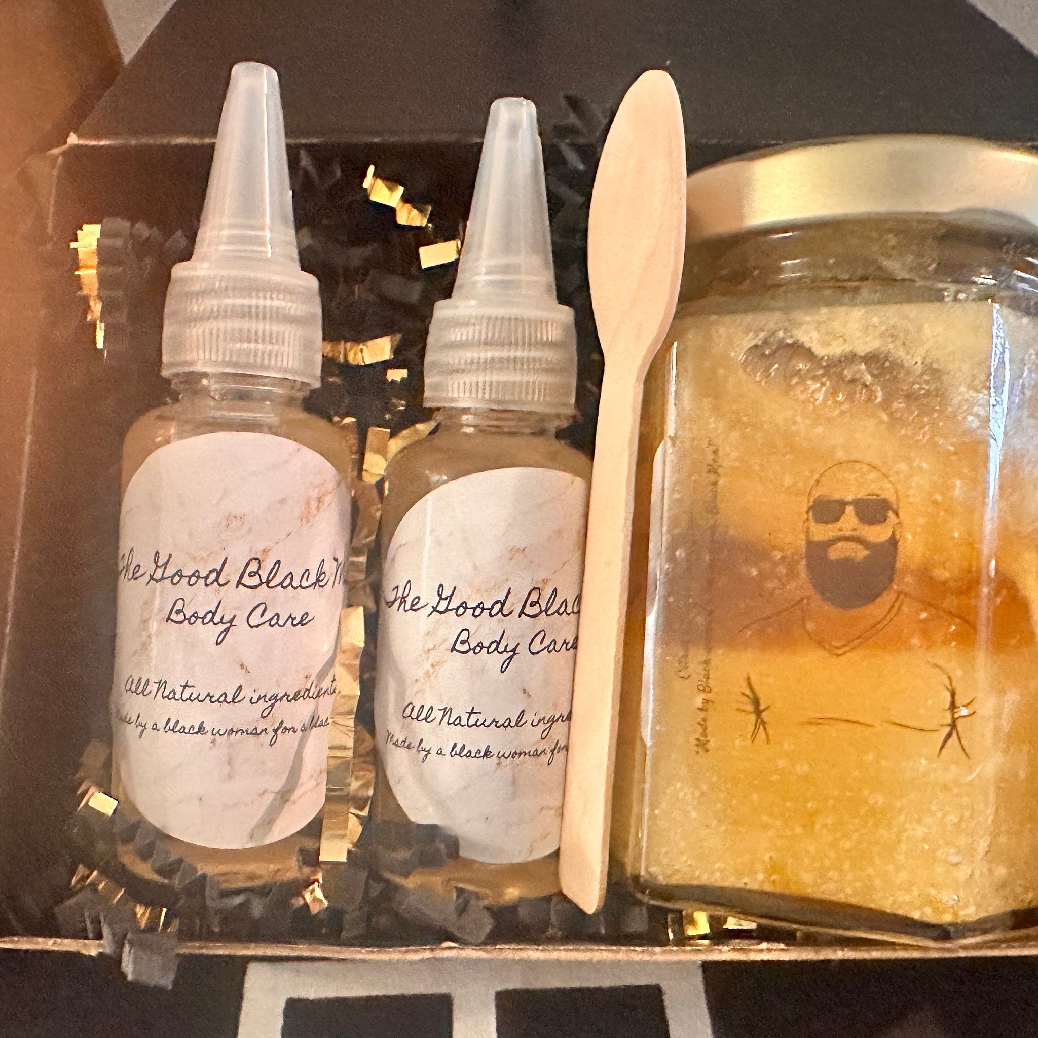 Body care set with clear bottles, wooden spoon, and jar with "The Good Black" text