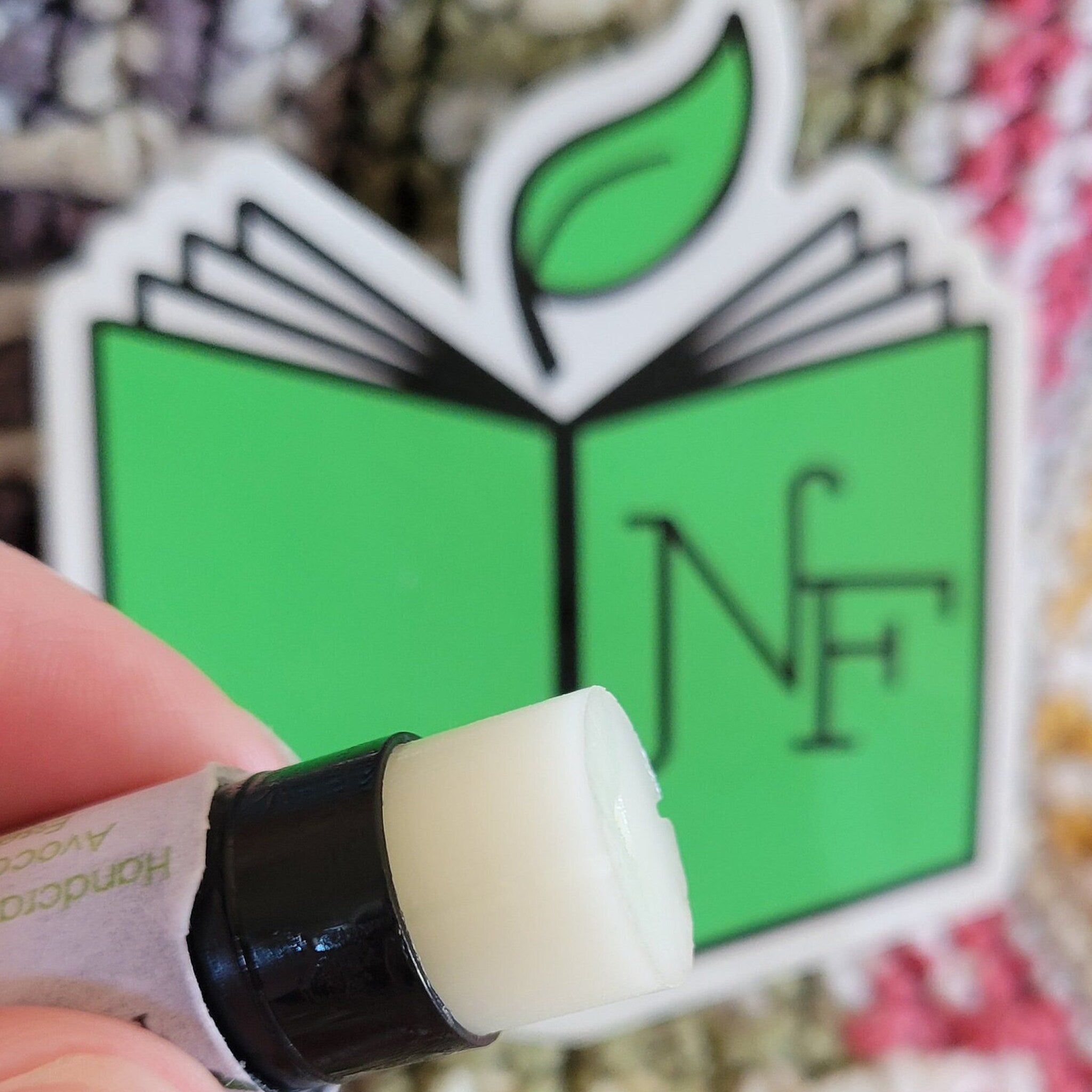 Lip balm stick with white balm, black tube, and paper label, with green logo in background