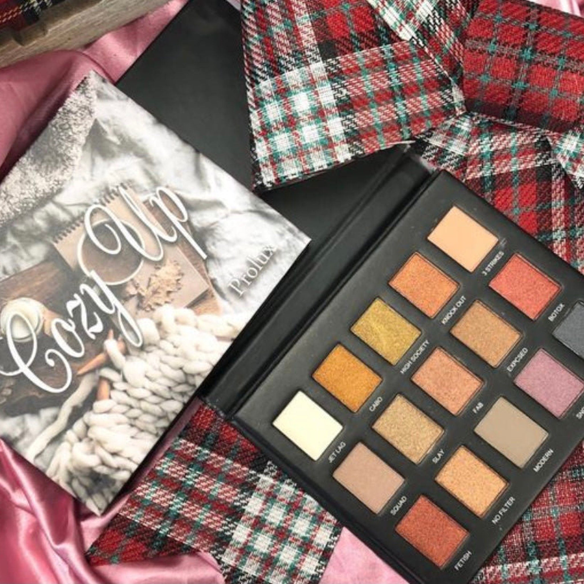 Eyeshadow palette with multiple colors in black case, plaid fabric and "Cozy Up" cover art