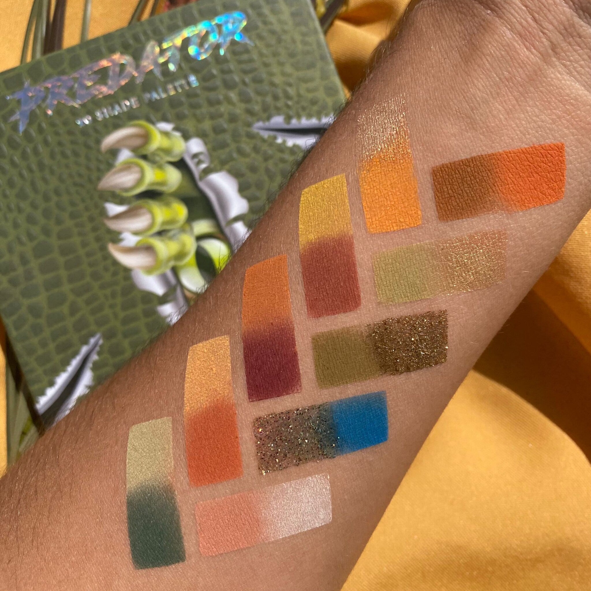 Eyeshadow swatches with various colors, including glitter, on arm with green textured palette in background