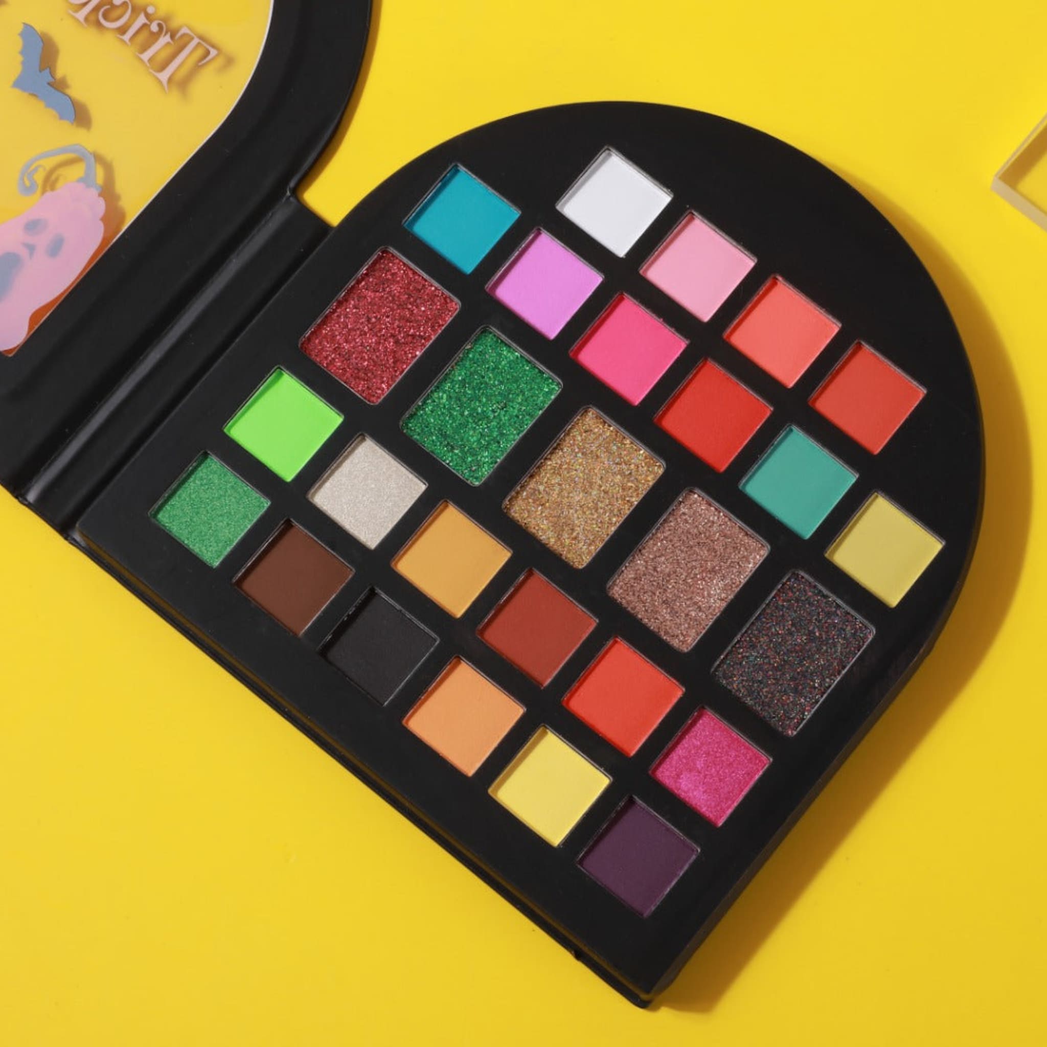 Eyeshadow palette with multiple colors, glitter, and matte shades in a black case on yellow background