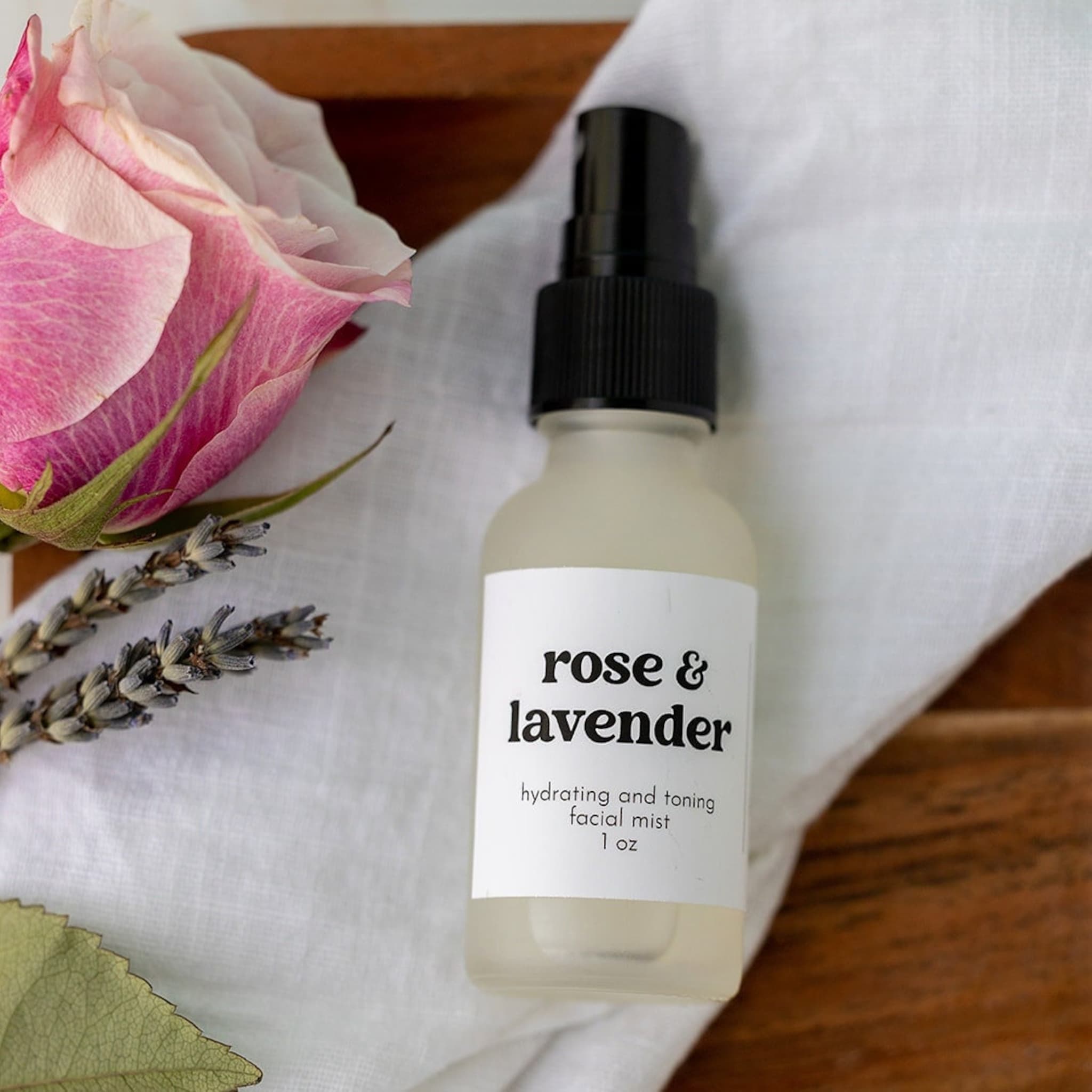 Facial mist bottle with white label, black cap, and "rose & lavender" text on white fabric