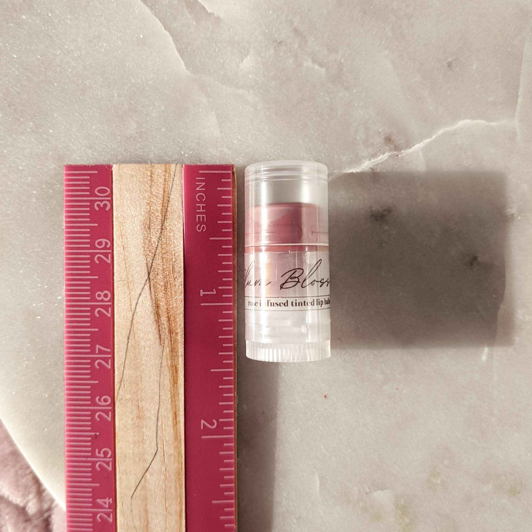 Lip balm with clear plastic tube, pink tinted color, and "Blossoms" text label next to a ruler
