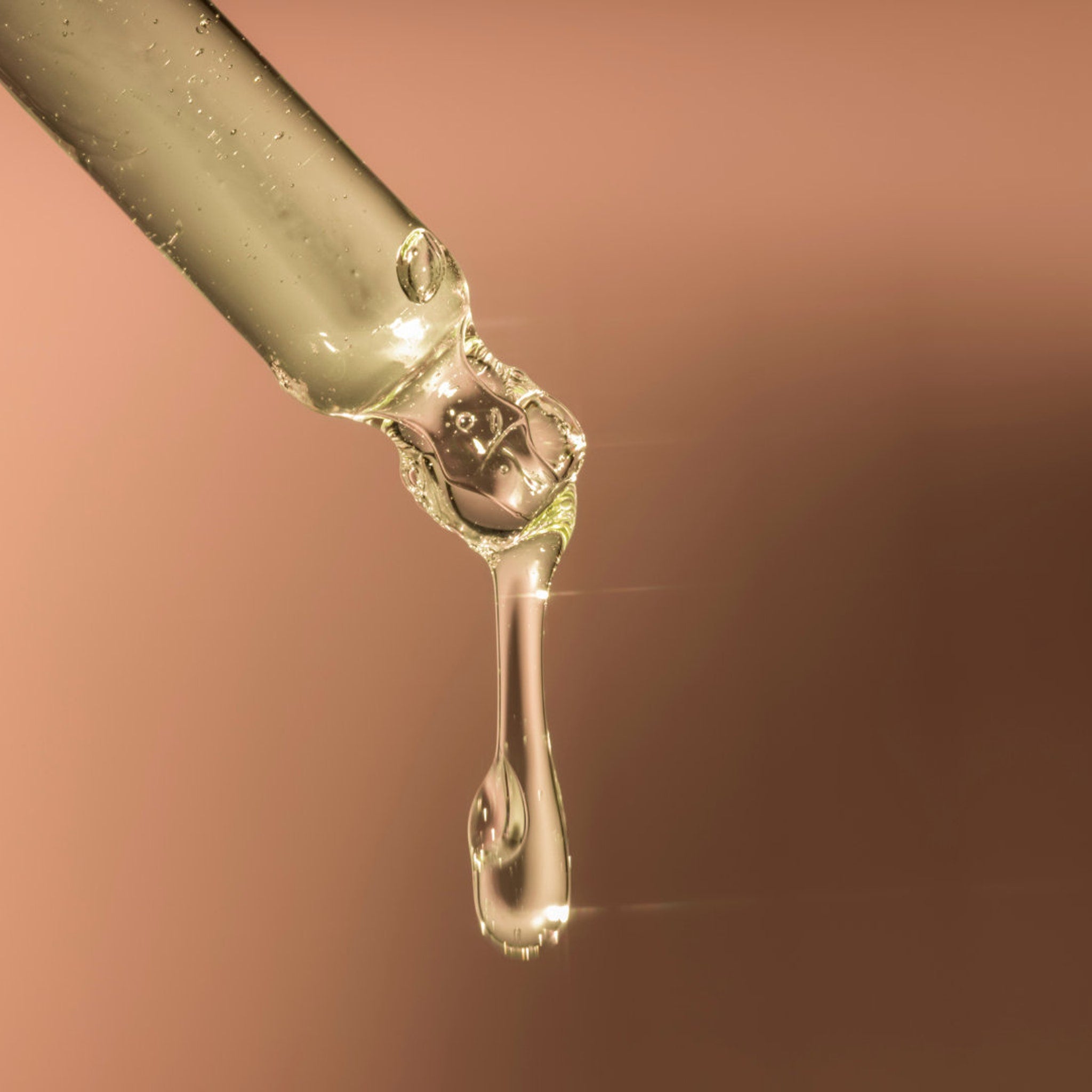 Dropper with clear liquid, glass, and a single drop falling against a brown background