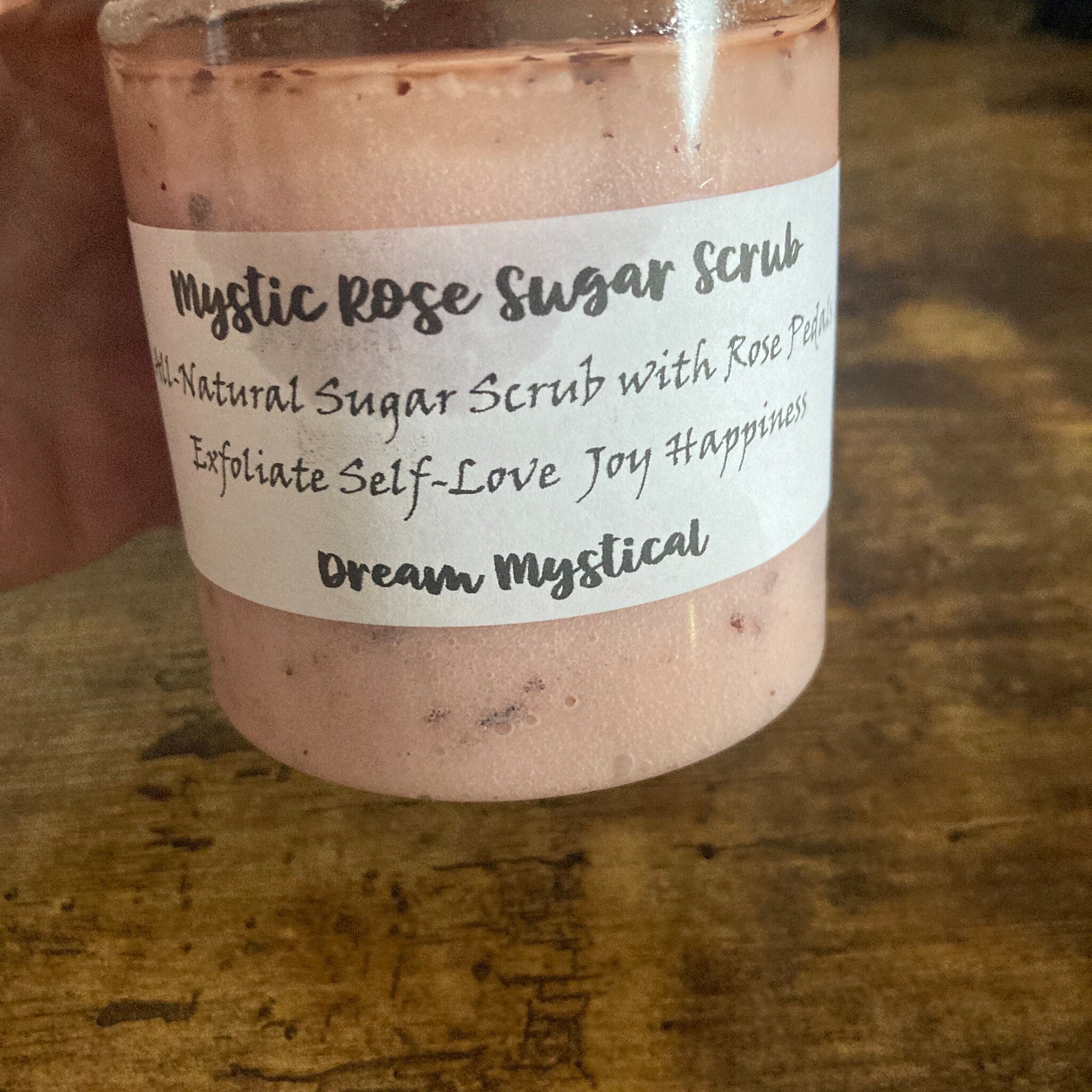 Sugar scrub in a pink jar with white label, "Mystic Rose" text, on wooden surface
