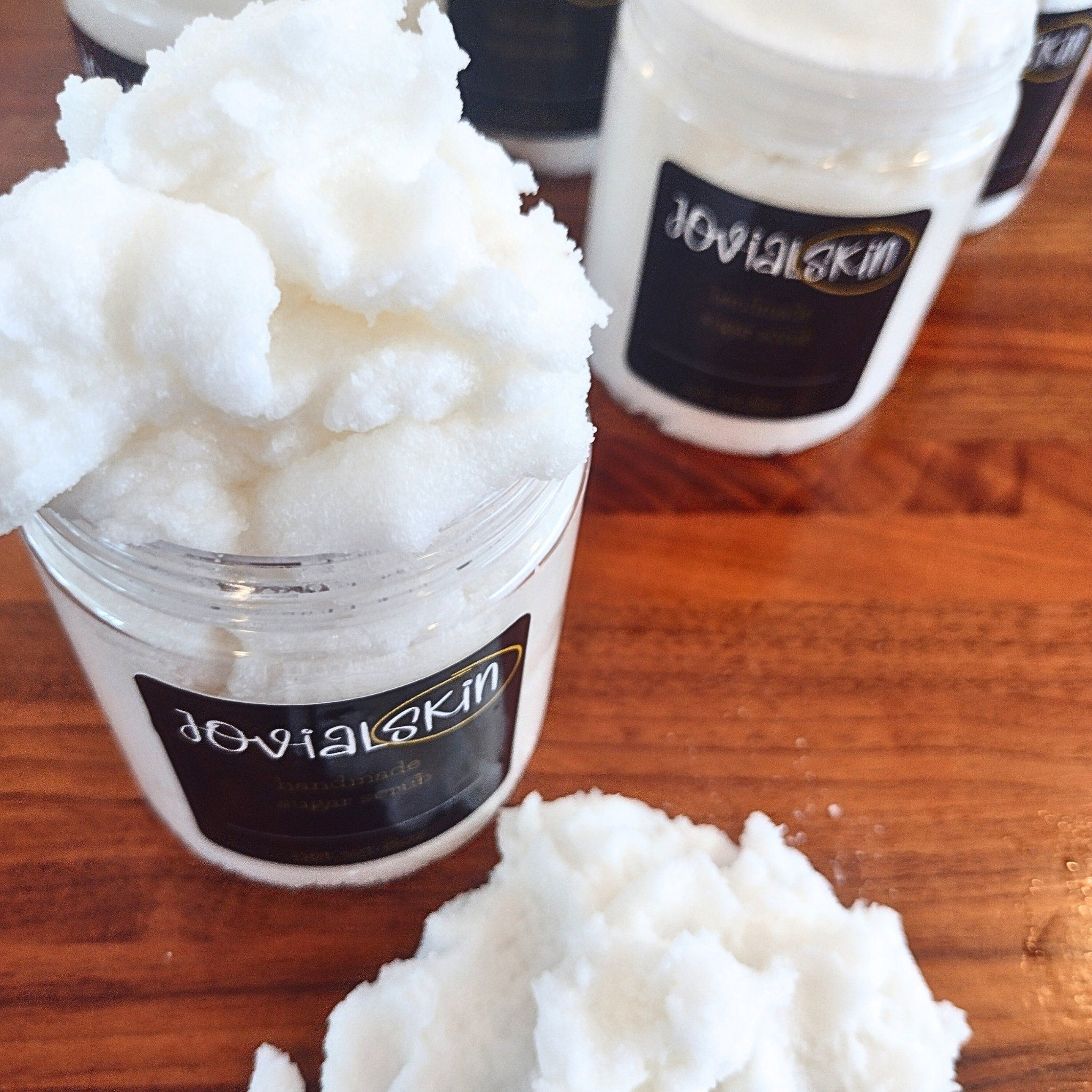 Sugar scrub in clear jar with white scrub, Jovial Skin label on wooden surface