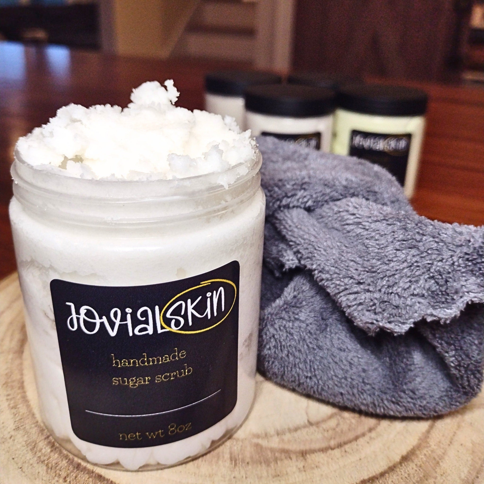 Sugar scrub in a clear glass jar with white contents, a black label, and a gray washcloth next to it