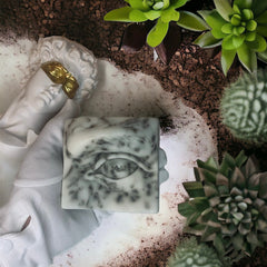 Soap bar with eye design, white and grey marble pattern, held by gloved hand near plants