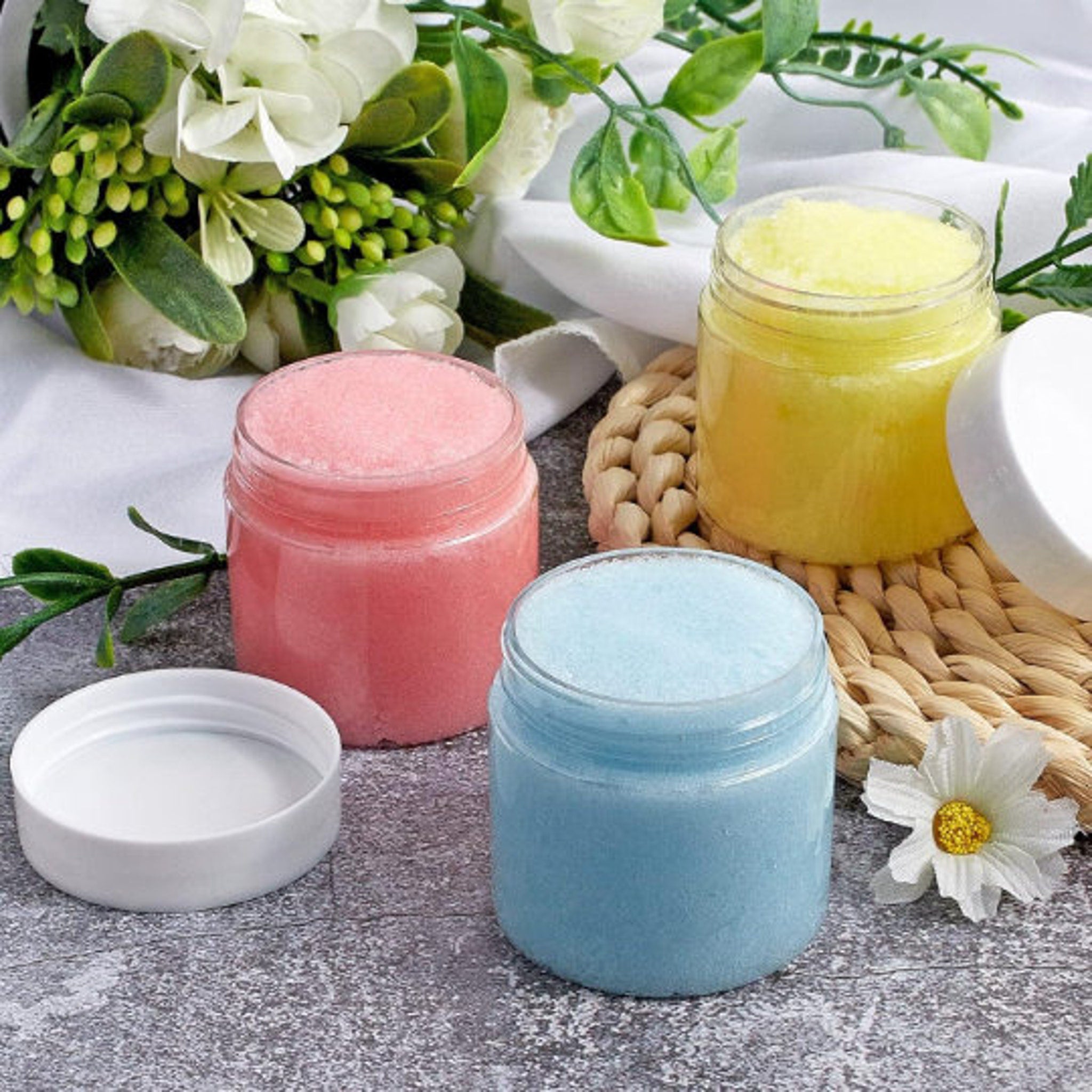Sugar scrub samples in clear jars with pink, yellow, and blue scrub, on woven mat
