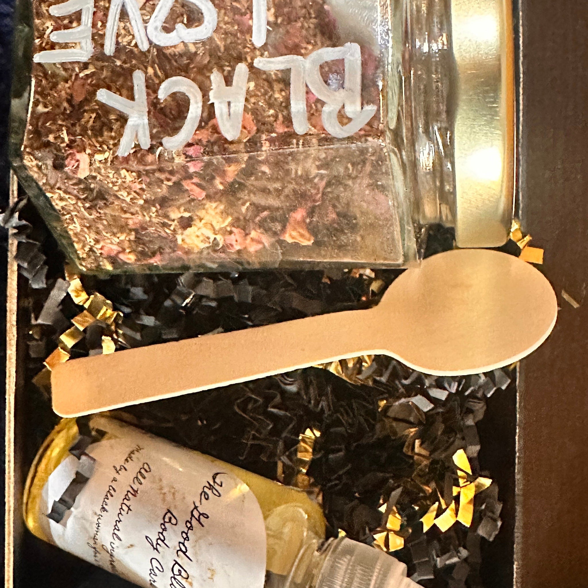 Black Love jar with white lettering, wooden spoon, and black and gold shredded paper in box