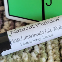 Lip balm tube with white label, black text, and Natural Fictions brand name on a textured background