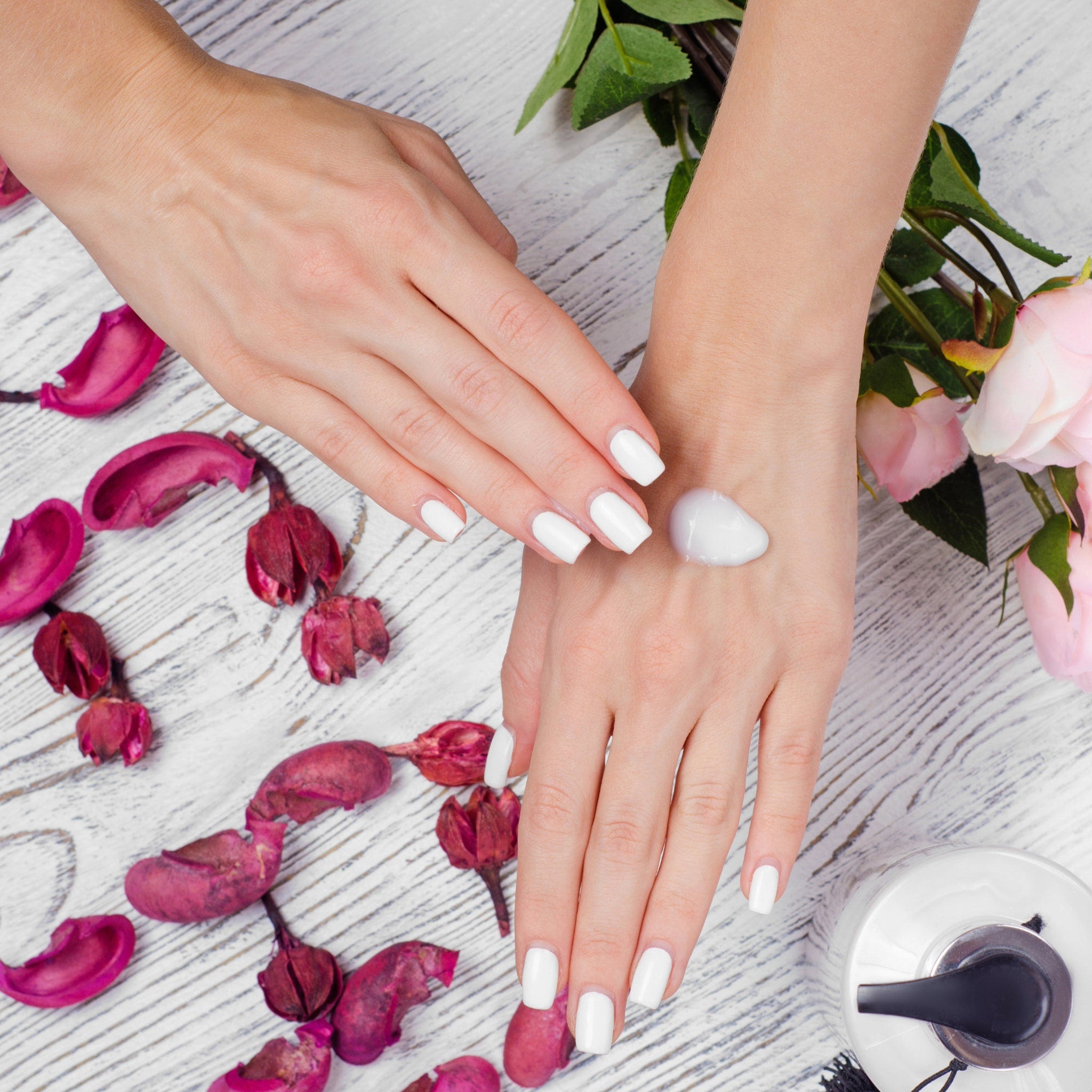 Manicure with white nails and cream, on a wood table with pink flowers and petals