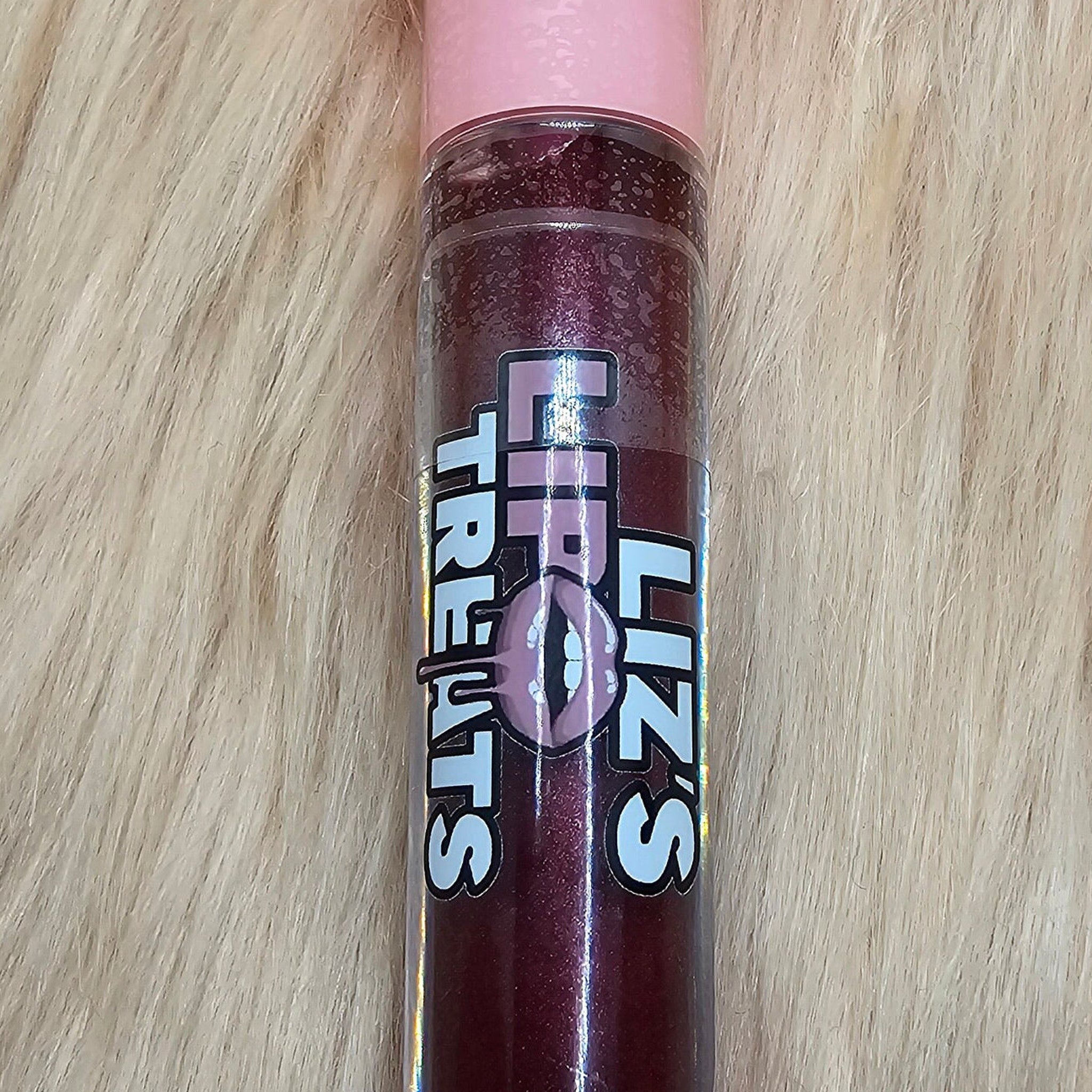 Lip gloss tube with pink cap, dark red shimmer, and Liz's Treats logo on a fur background