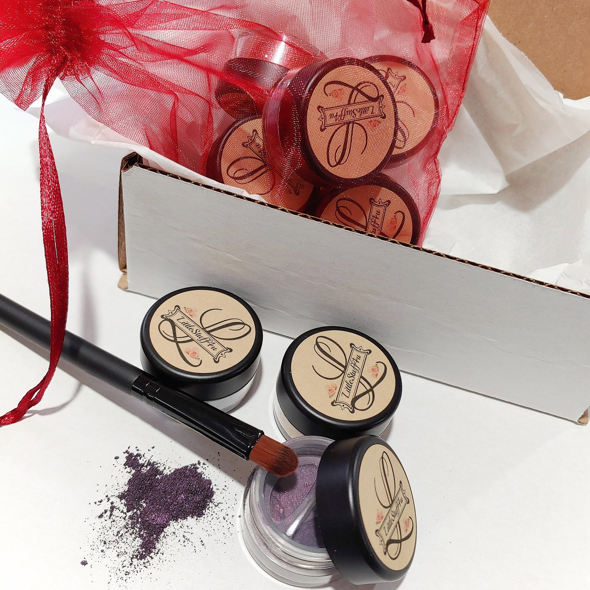 Eye shadow pots with black lids, purple powder, and Little Stuff4u branding, with a brush