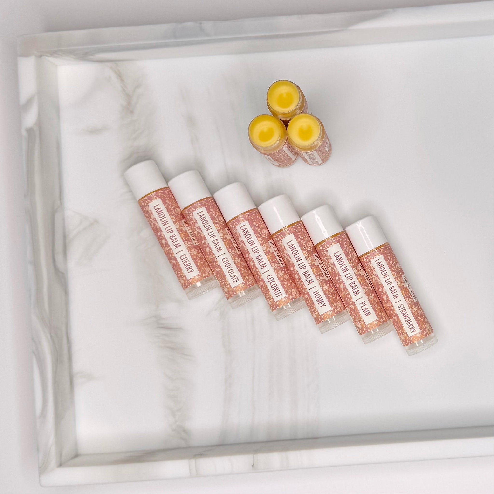 Lanolin lip balm tubes with white caps and pink glitter label, on marble tray, Hue & Lather brand