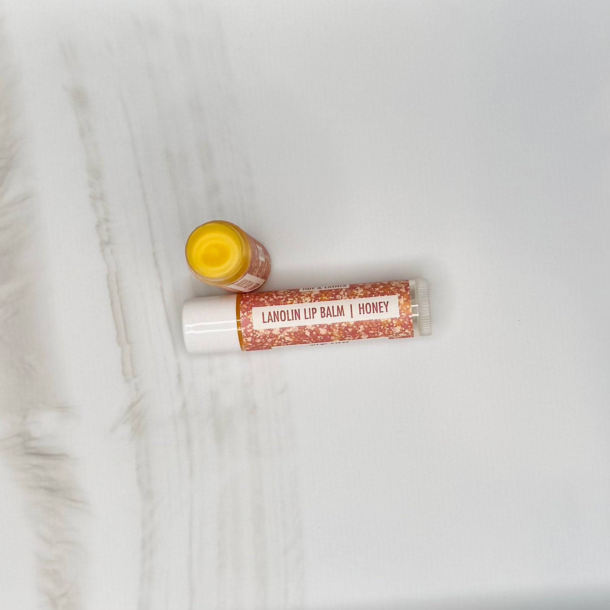 Lanolin lip balm, white tube with red speckled label, yellow balm visible, Hue & Lather brand