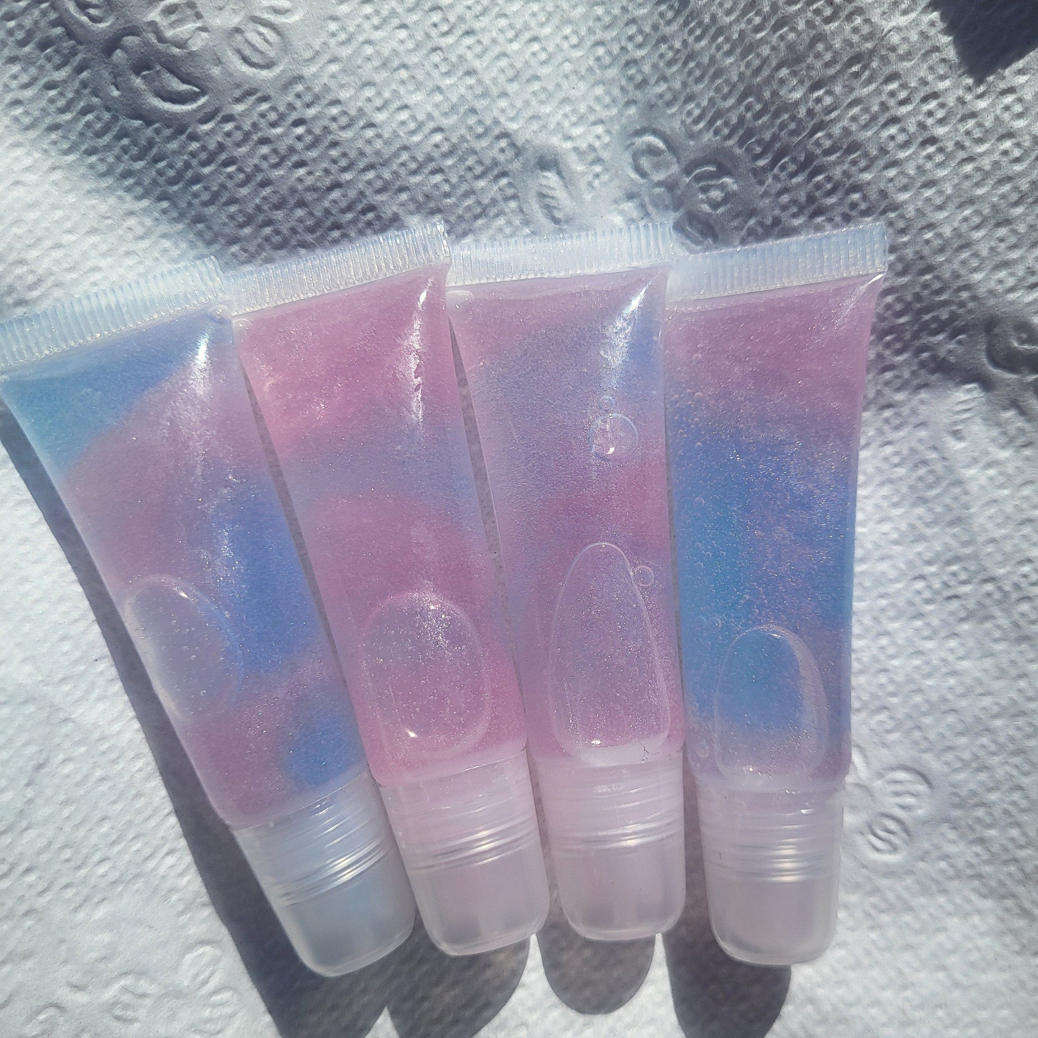 Lip gloss tubes with pink and blue iridescent color, clear plastic tubes, on white textured surface