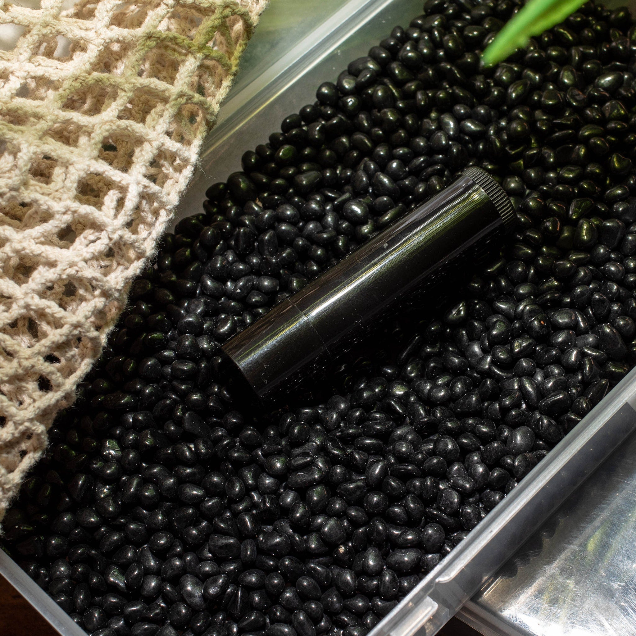 Black lipstick tube on black pebbles next to beige woven net bag