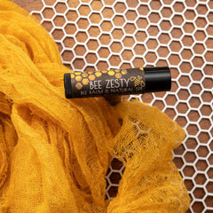 Lip balm tube with "Bee Zesty" label, black tube, yellow fabric, and honeycomb background.