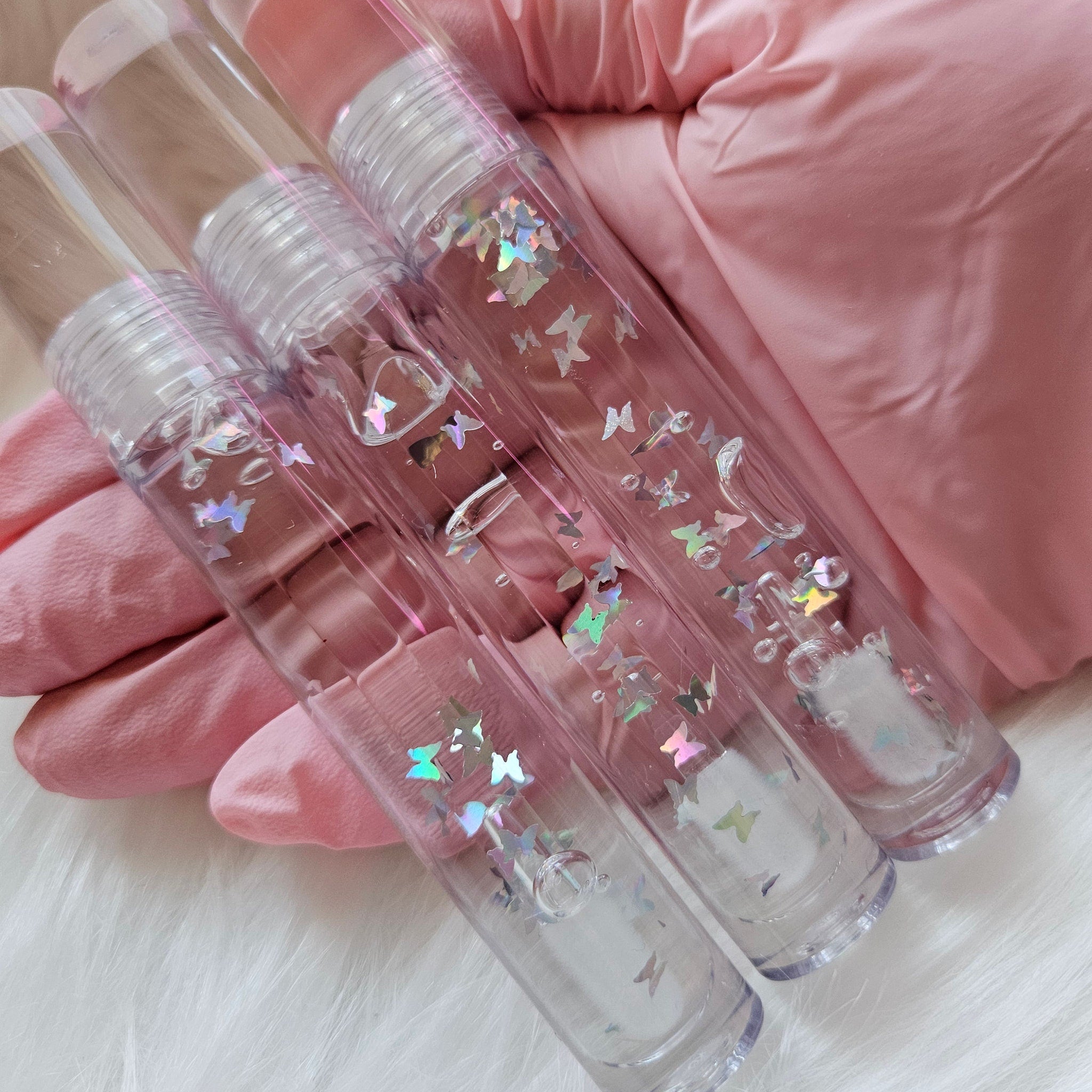 Lip gloss tubes with clear plastic, butterfly glitter, and pink lid, held in gloved hand.