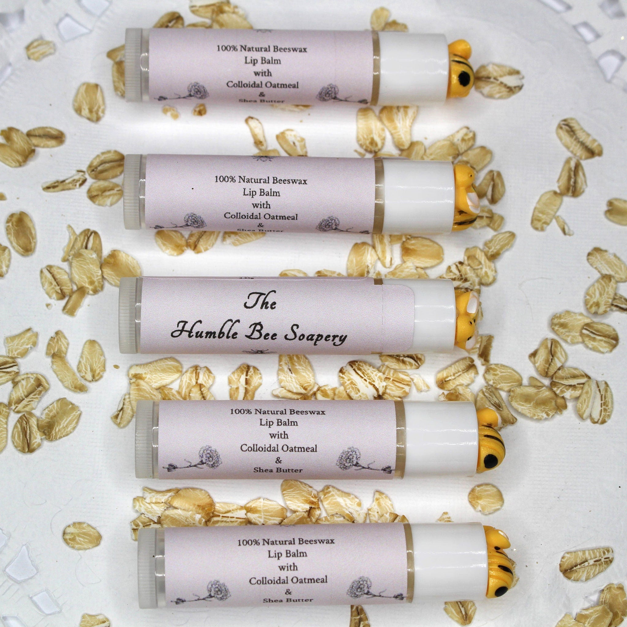 Lip balm tubes with white and lavender labels, yellow bee ends, and oats scattered around them
