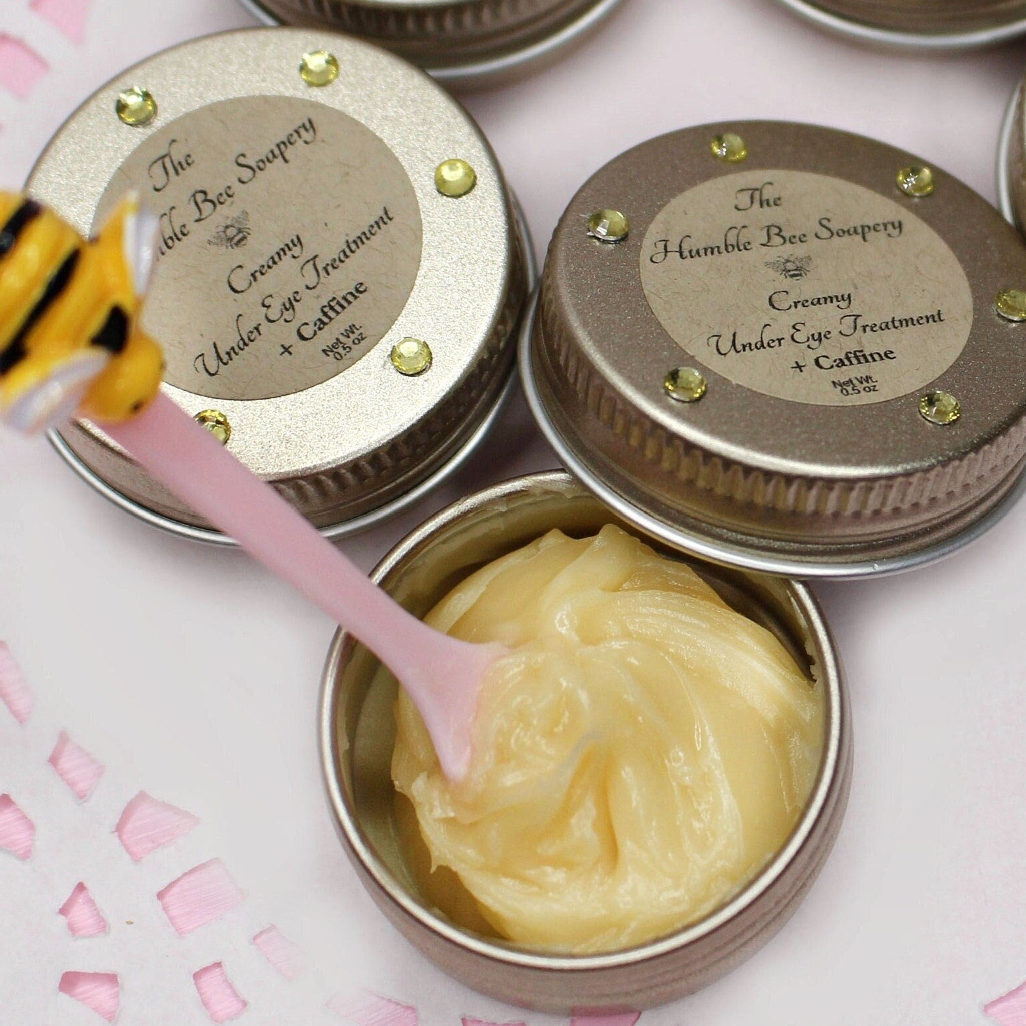 Under eye treatment cream in a silver tin with a pink scoop, The Humble Bee Soapery label visible