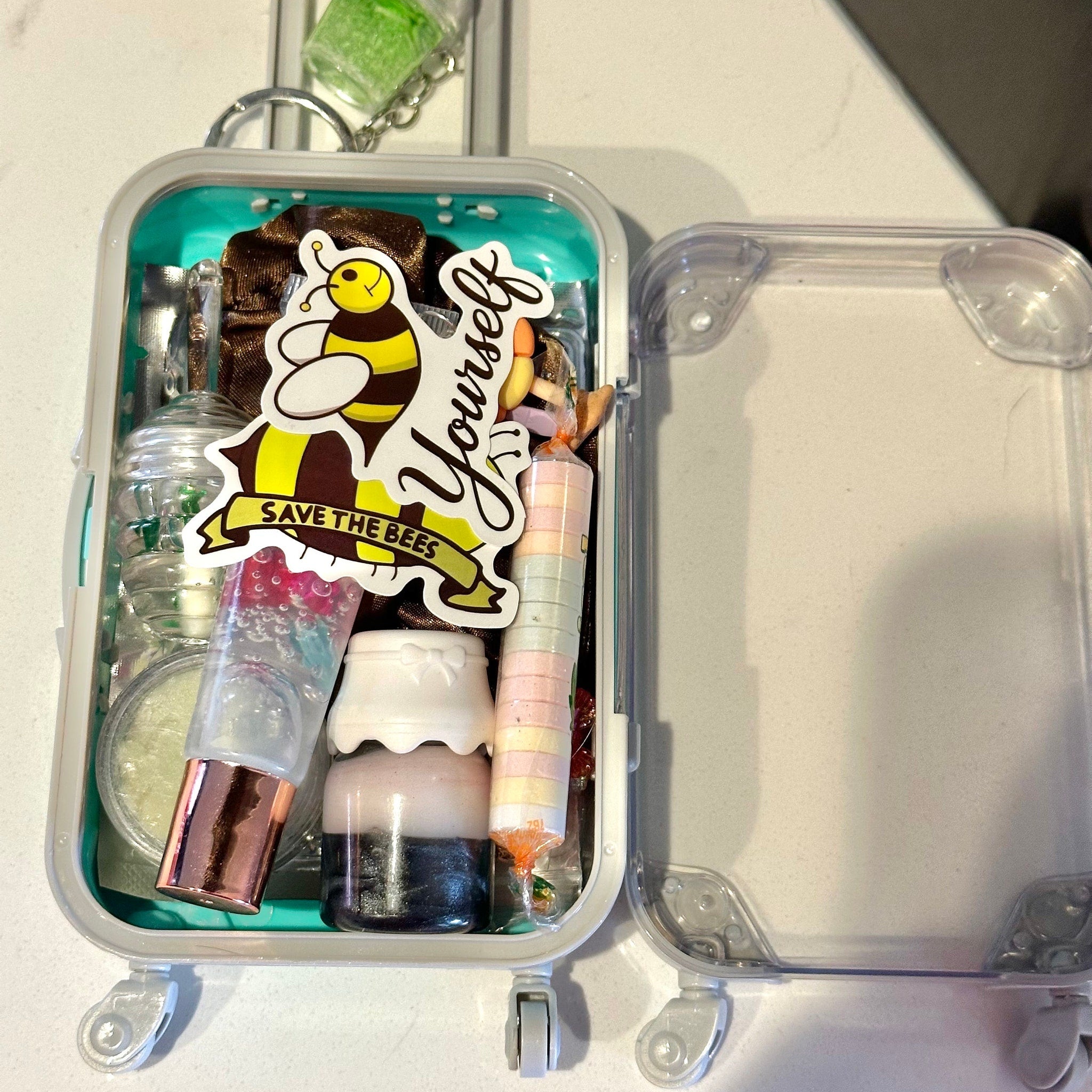 Miniature suitcase with teal interior, clear plastic exterior, and Save the Bees sticker inside
