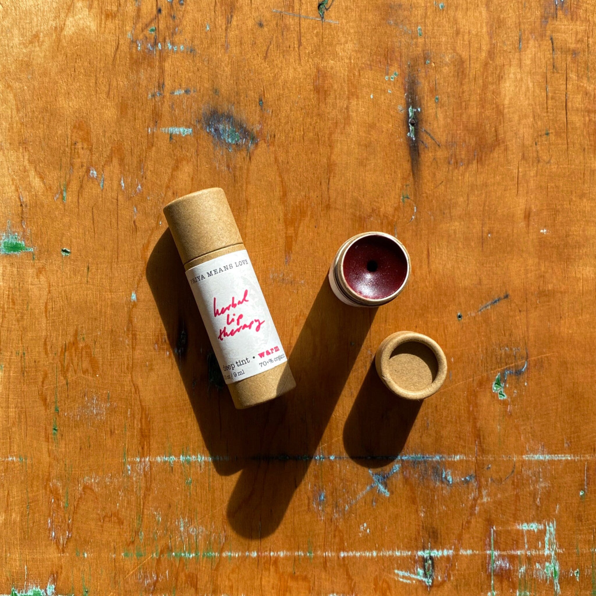 Lip balm tube with brown paper packaging, deep red tint, and open lid on wooden surface
