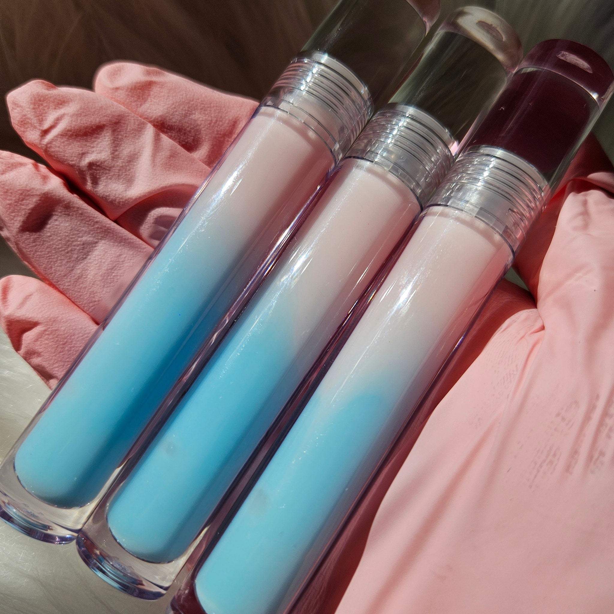 Lip gloss tubes with blue, pink, and clear gradient colors held in pink gloved hand