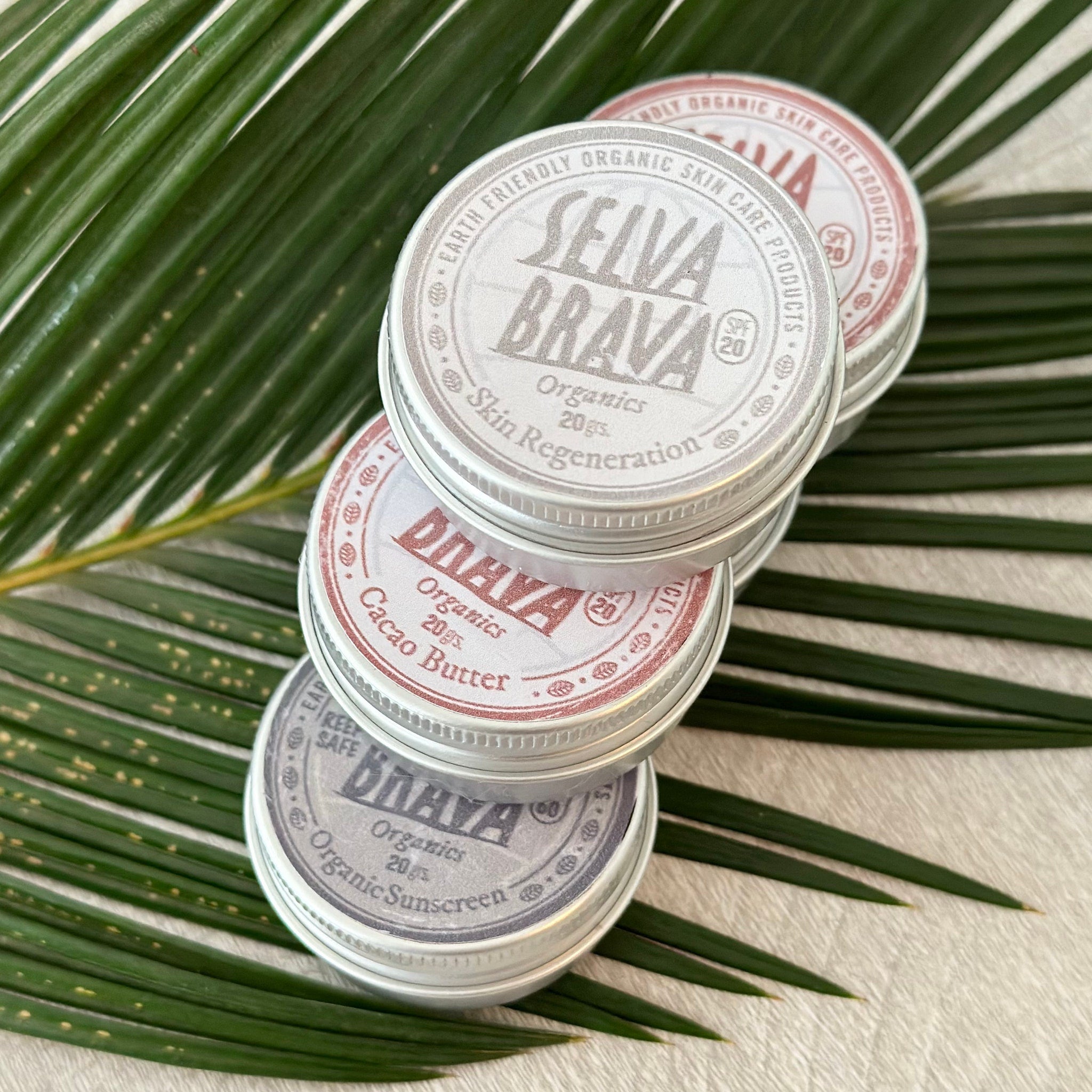 Organic skin care products in silver tins, stacked with visible brand names, on green leaves