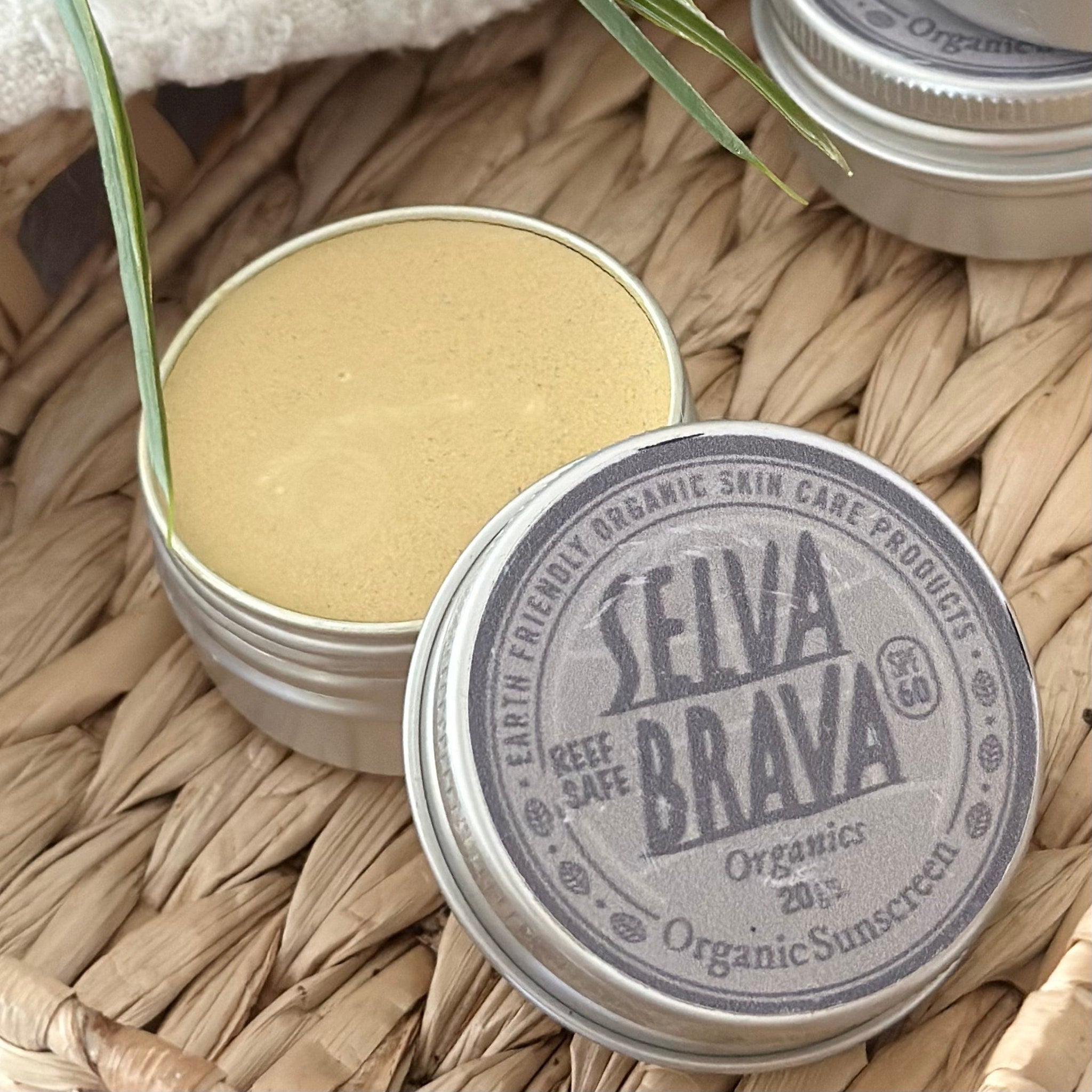 Organic sunscreen in silver tin with tan cream, SELVA BRAVA branding, on woven surface
