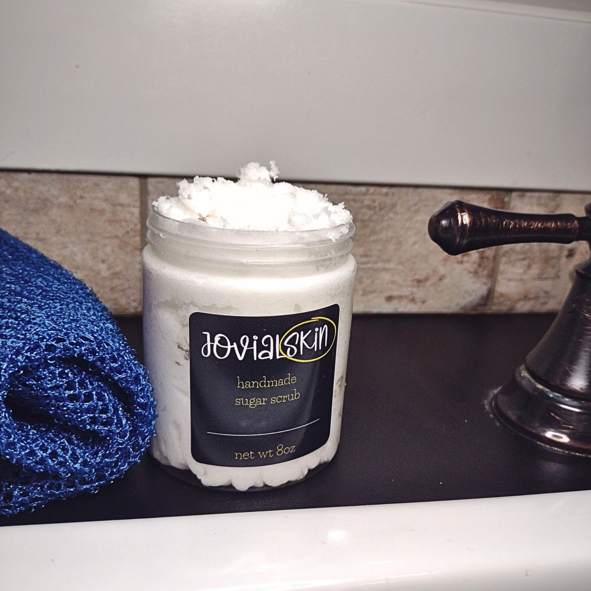 Sugar scrub in a glass jar with white contents and JovialSkin label next to blue washcloth