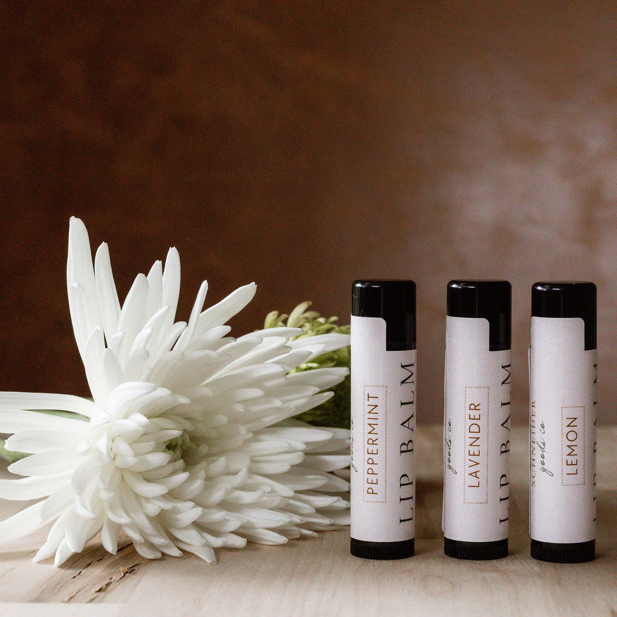 Lip balm tubes with white labels, black caps, and text, next to white flower on wood surface
