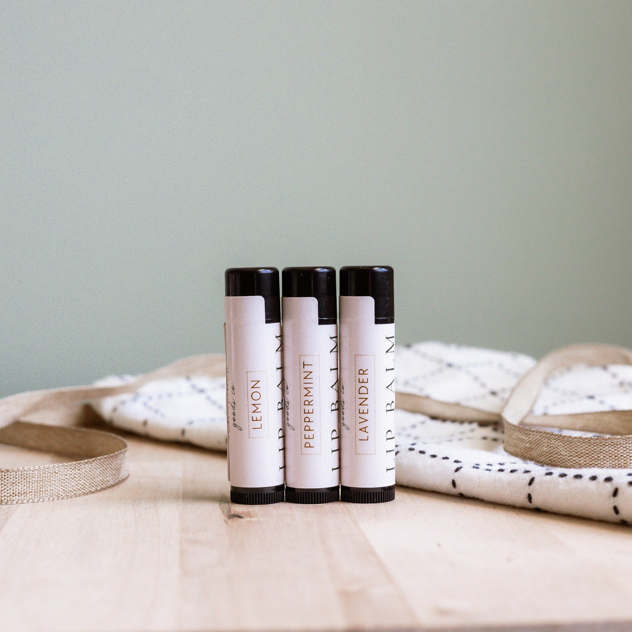 Lip balm tubes with black caps and white labels, on a wooden surface, with text "Lemon", "Peppermint", "Lavender