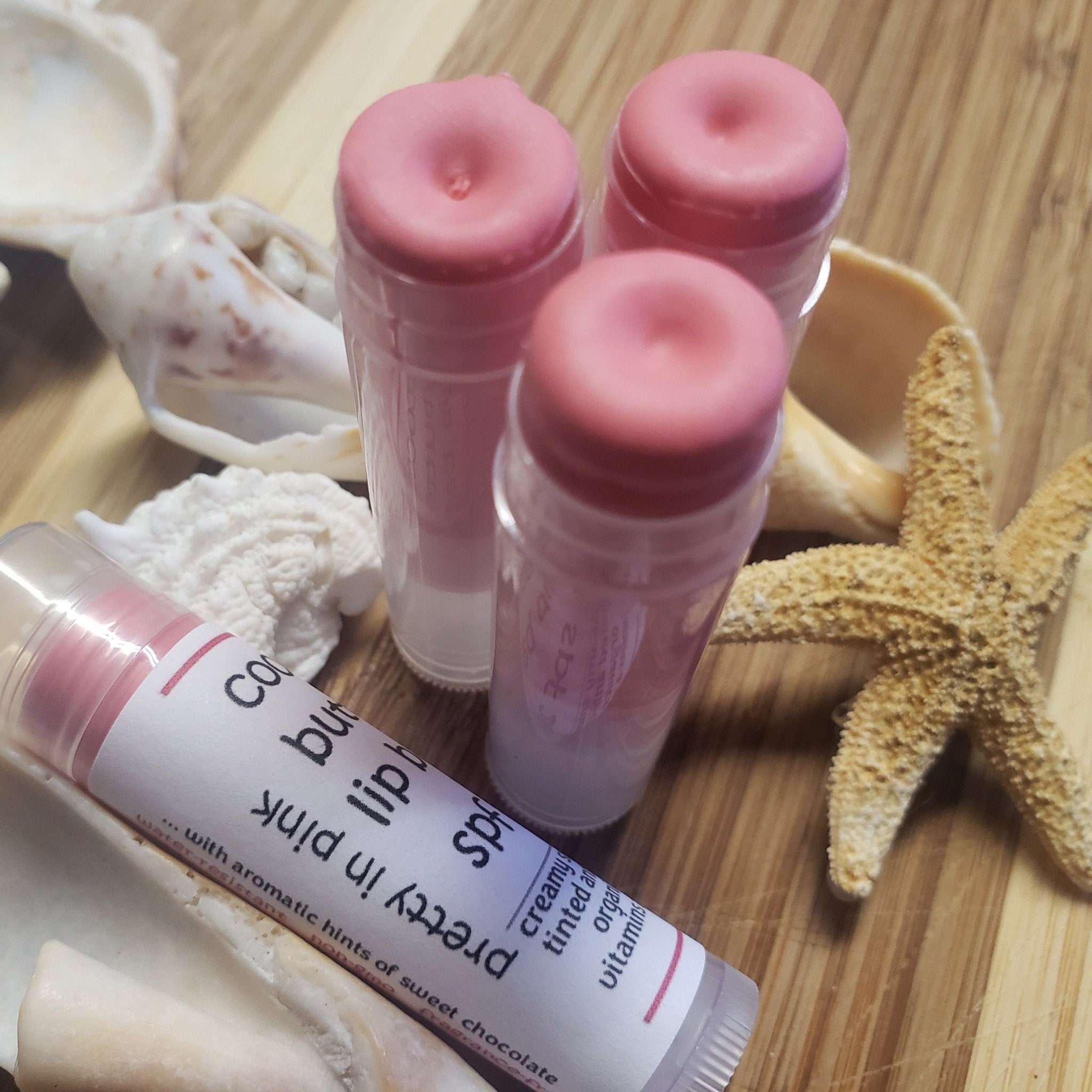 Lip balm tubes with pink tops and clear tubes, displayed with seashells and starfish on wood