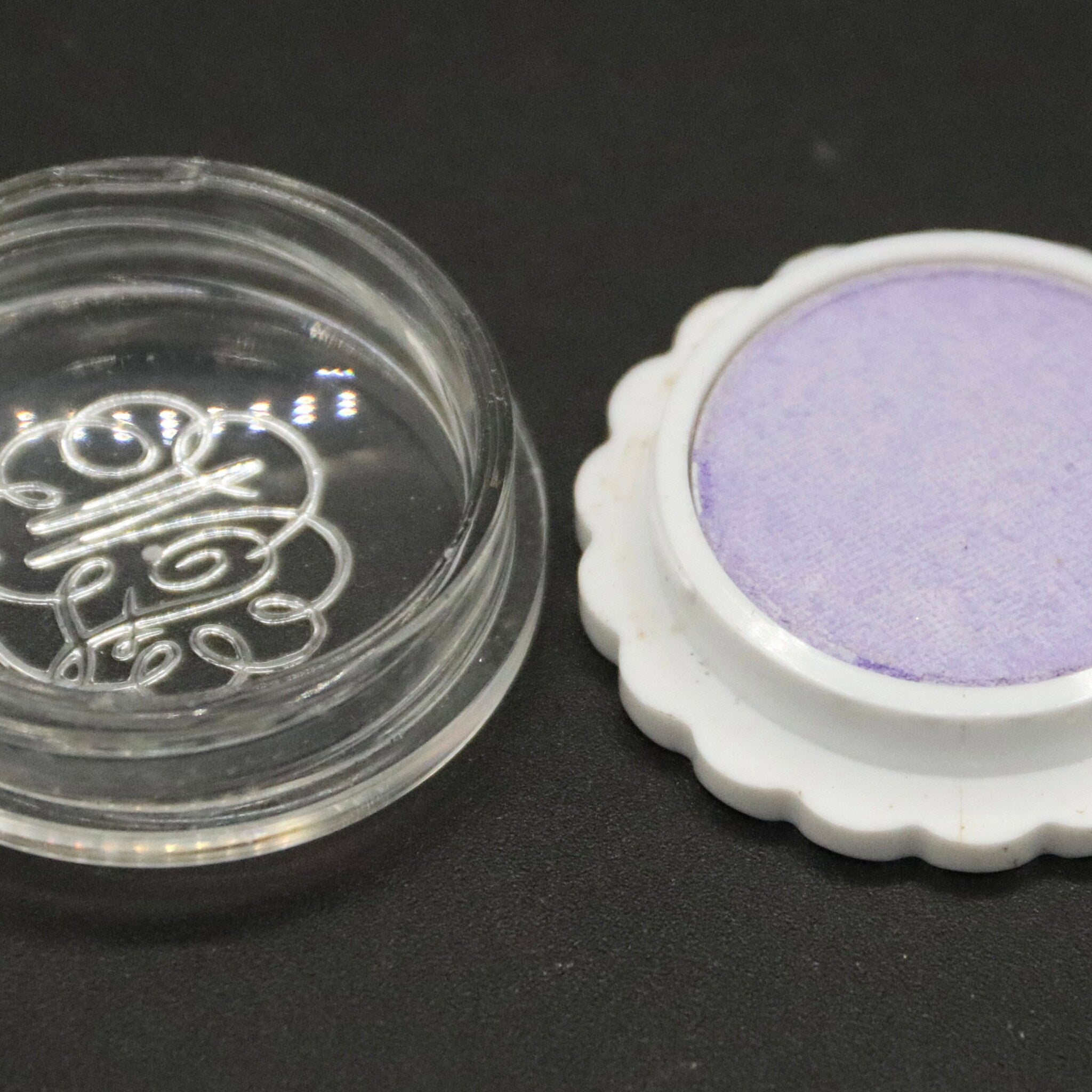 Purple eyeshadow compact with white scalloped edge and clear container with logo detail