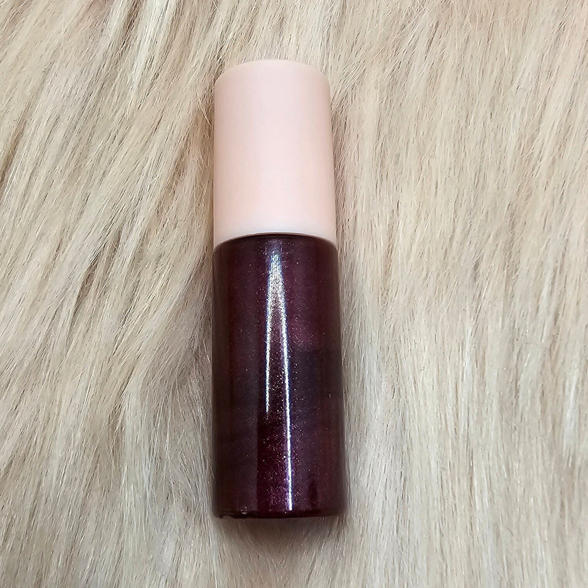 Lip gloss tube with pink cap, dark purple gloss, and reflective shine on a furry surface