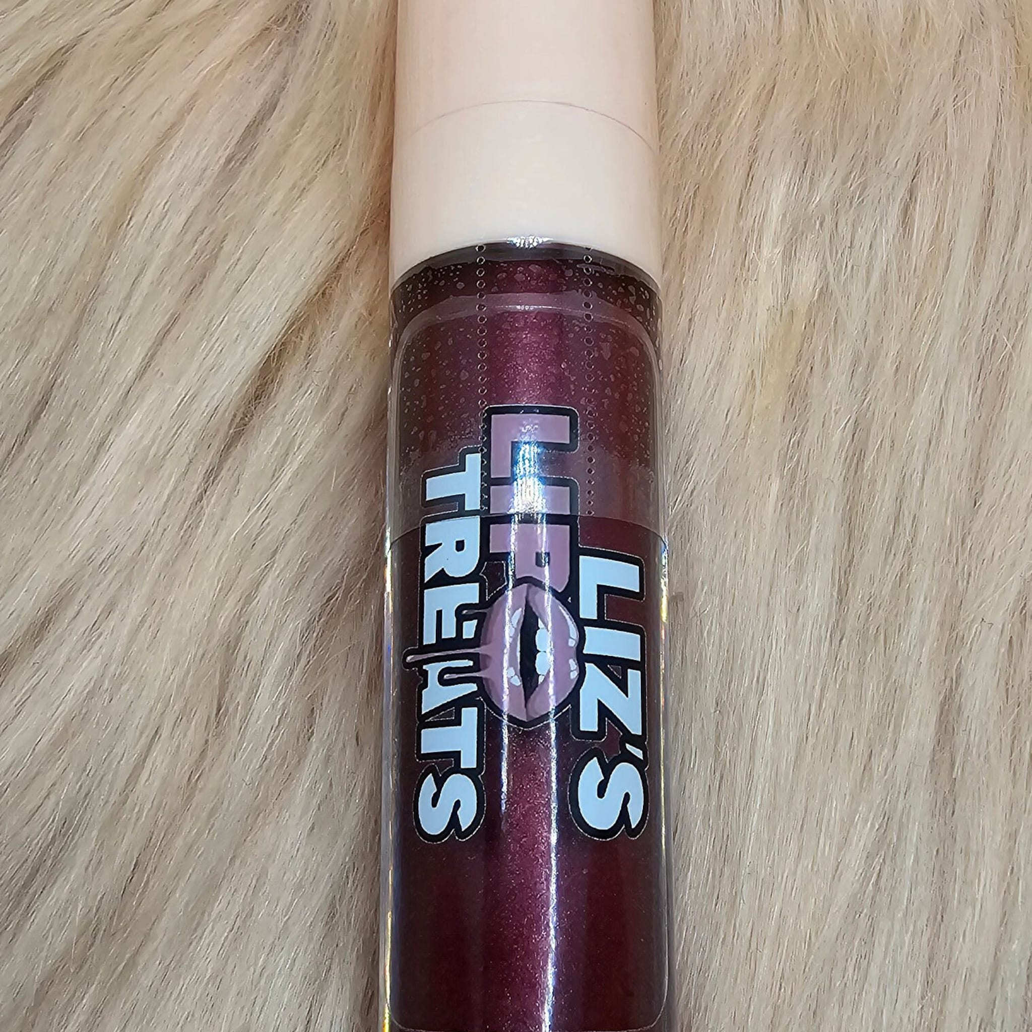 Lip gloss tube, dark red with glitter, white cap, "Liz's Treats" label on fur background