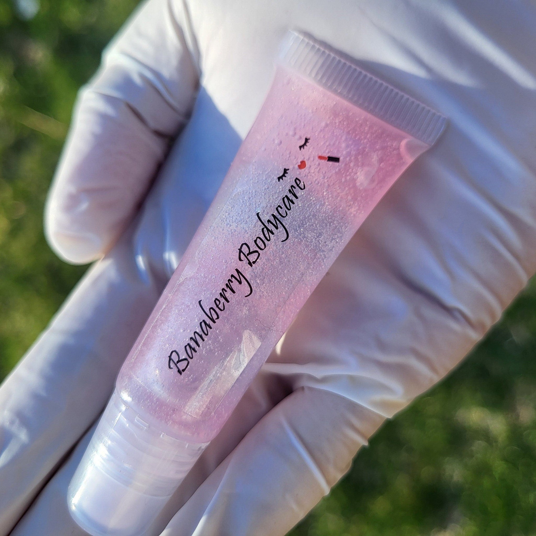 Lip gloss tube, pink transparent with glitter, held in a gloved hand, Banaberry Bodycare text visible