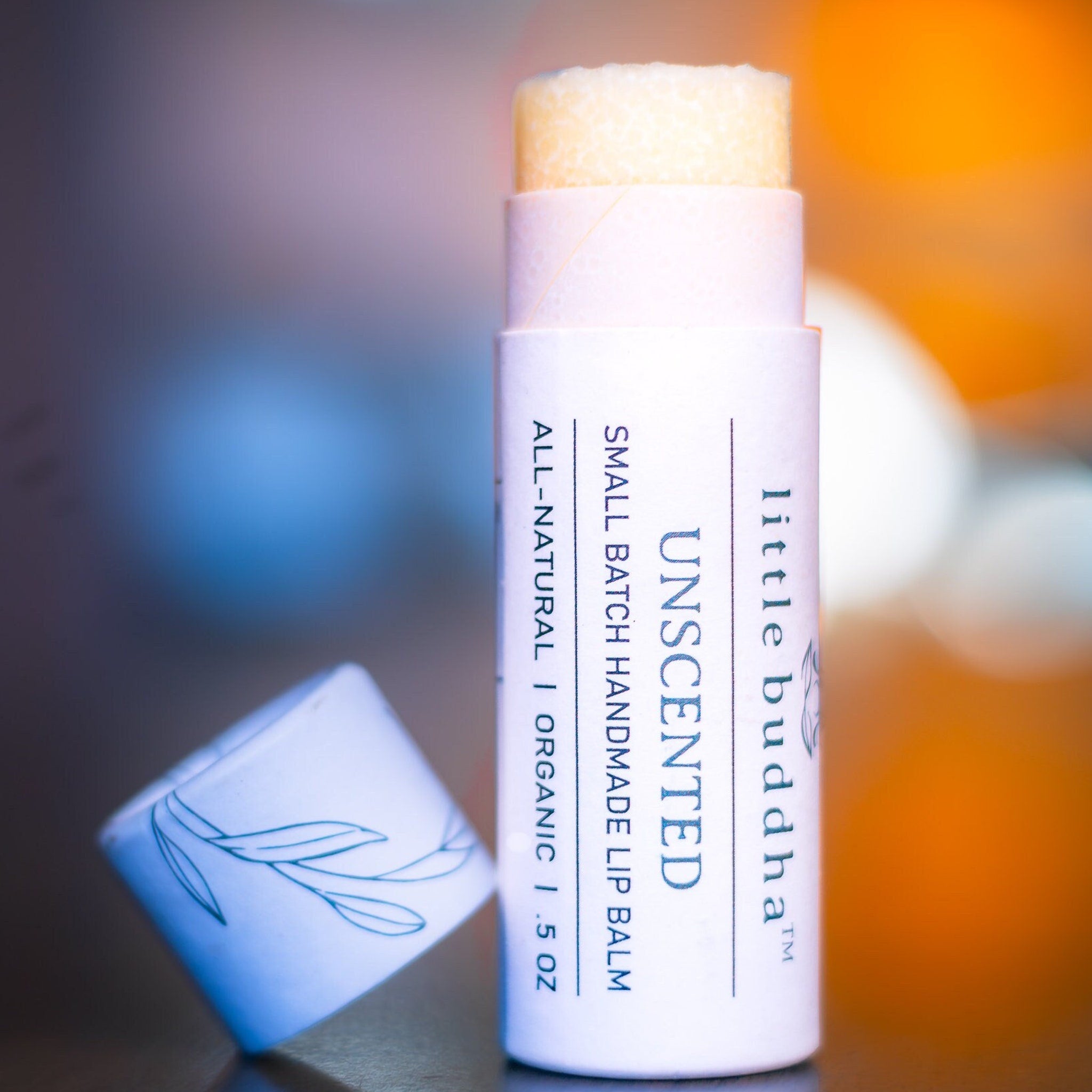 Lip balm stick with white and light pink tube, visible "Little Buddha" text, and exposed yellow balm