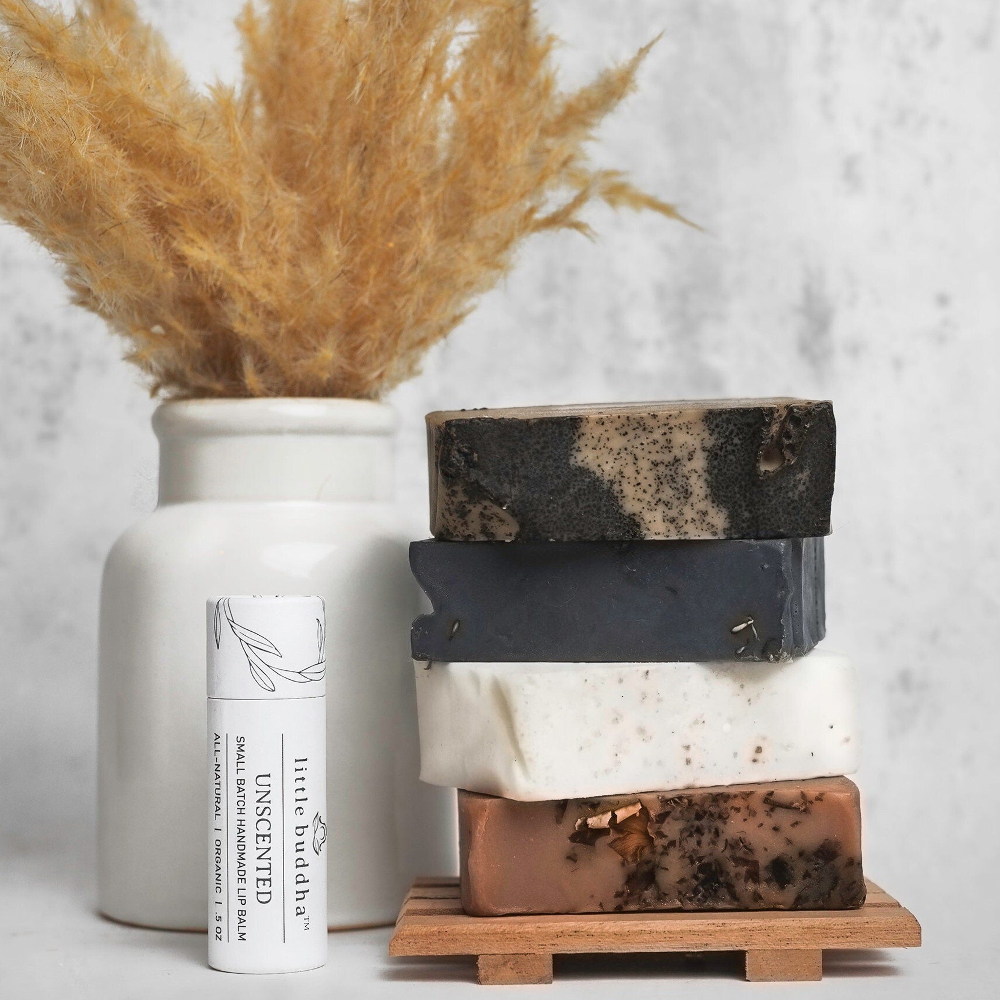 Soap bars stacked, various colors, with "little buddha" lip balm and dried flowers in vase