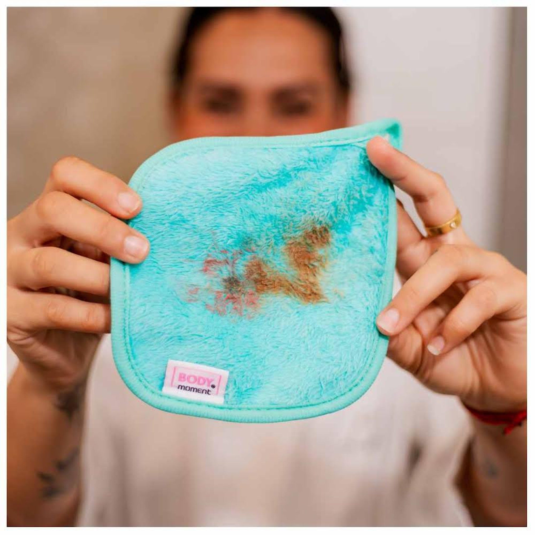 Makeup remover pad, turquoise plush material with stain, held by hands, Body Moment label visible
